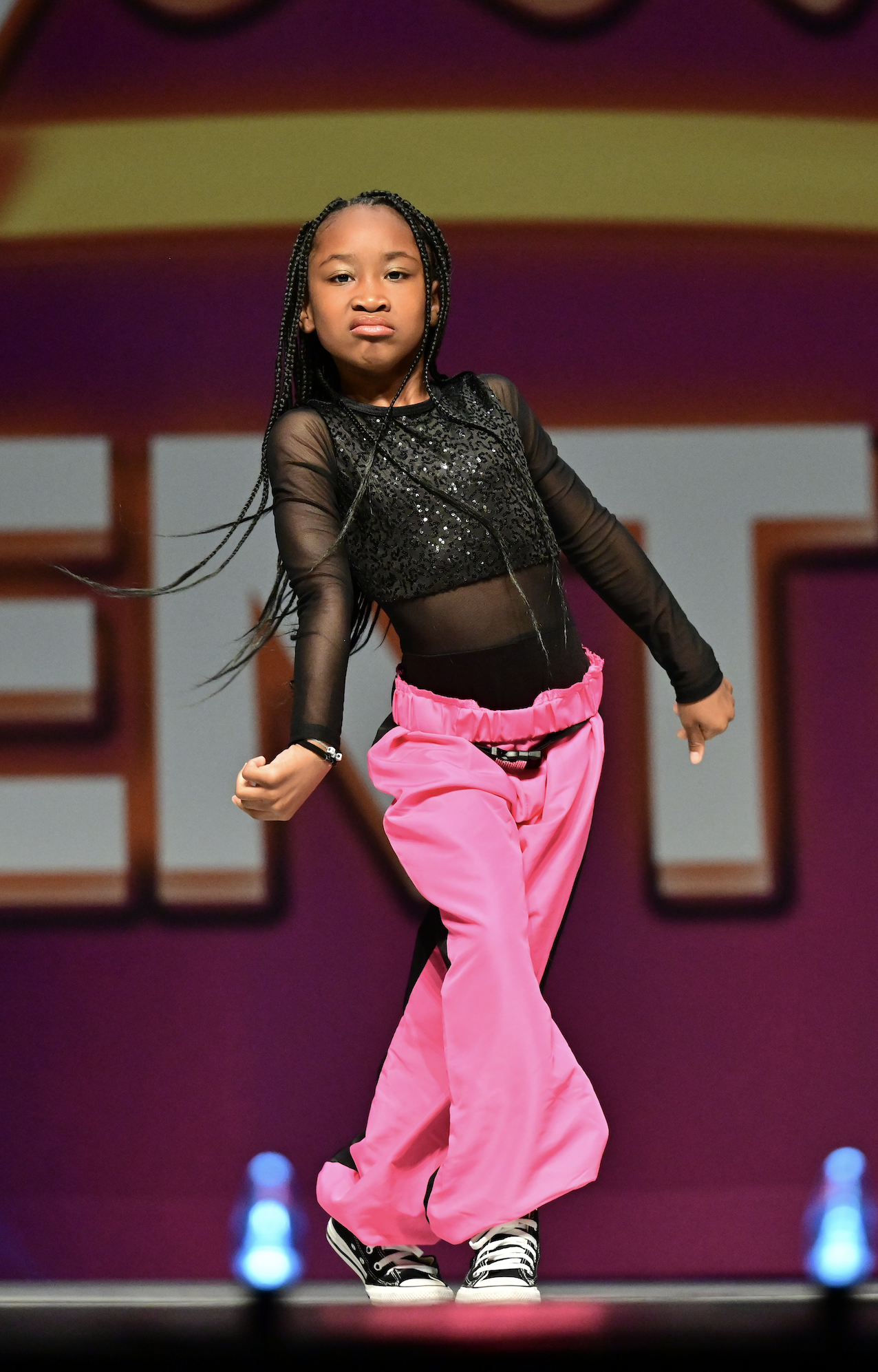 Young girl performing a dance or cheer move on stage, wearing black mesh top with sequins and bright pink pants, with braided hair and sneakers.