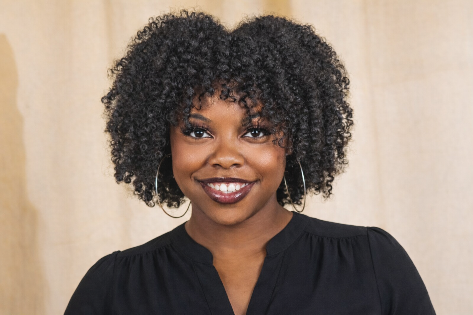 A smiling woman with dark skin and curly black hair, wearing hoop earrings and a black top, standing against a neutral background.