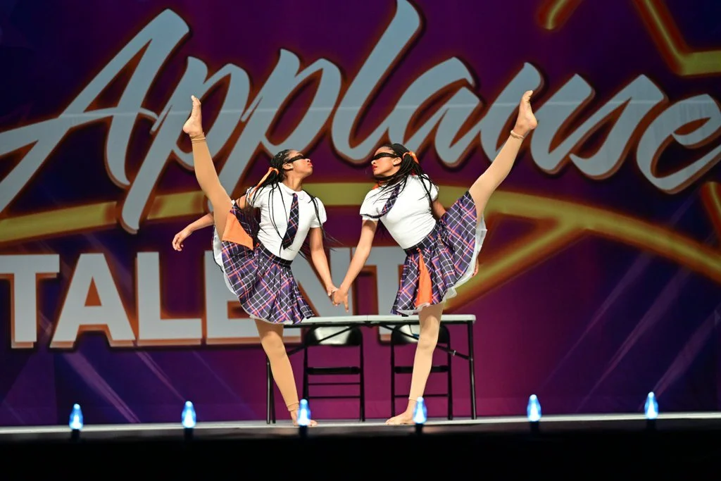 Two young girls in school uniforms performing a dance routine on stage, holding hands with their legs extended high in the air, in front of a large sign that says 'Apply to Dance'.