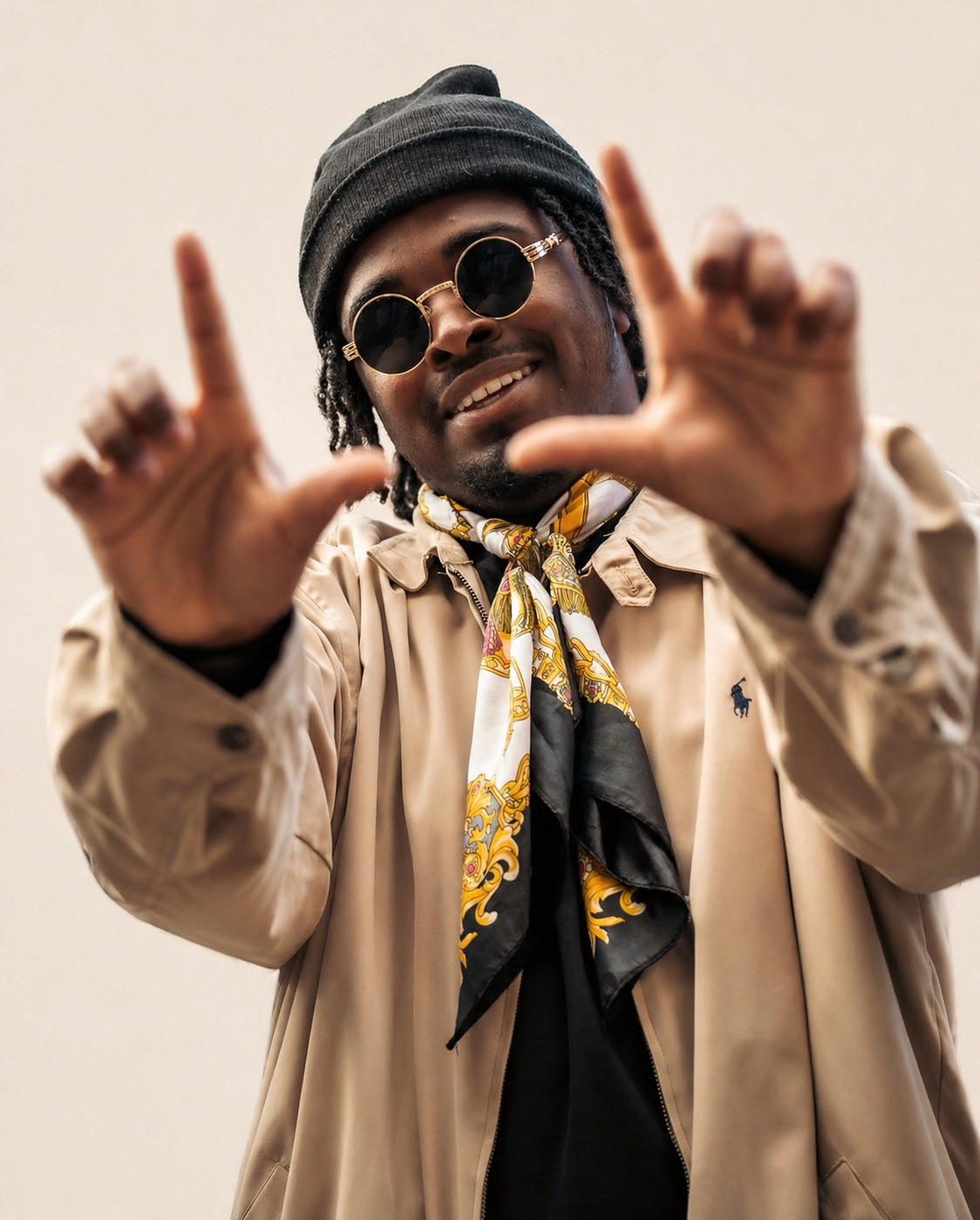 A person wearing round black sunglasses, a black beanie, a beige jacket, a colorful scarf, and making a framing gesture with their hands.