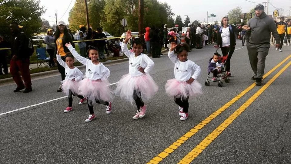 Four young girls dressed in white tops, pink tutus, and sneakers performing a dance routine on a street during a parade, with spectators and other participants in the background.