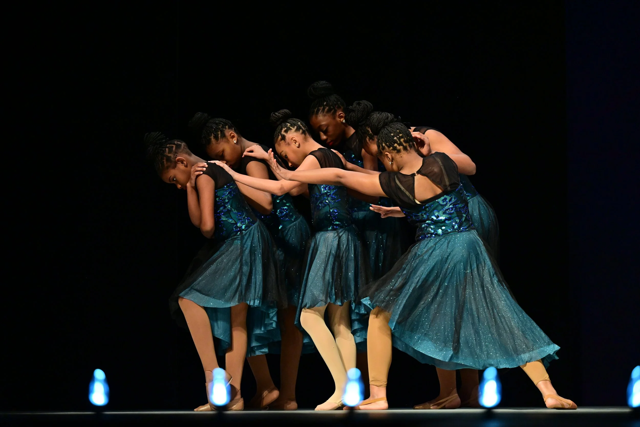 Six ballet dancers performing on stage in coordinated poses, wearing blue skirts and black tops, with dark background and stage lights.