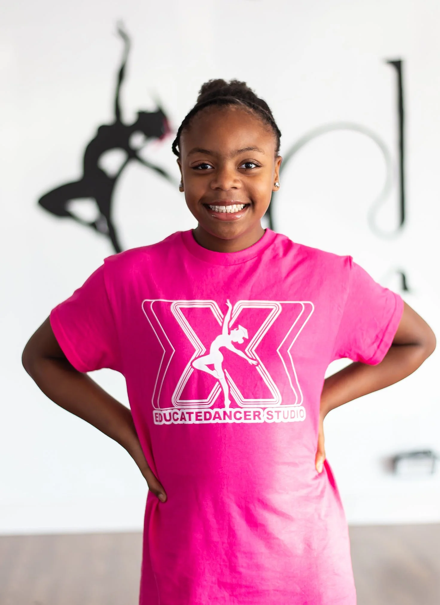Young girl with a bright smile wearing a pink T-shirt with a unicorn logo and the words 'Educate Dancer Studio,' standing with hands on hips in a dance studio.