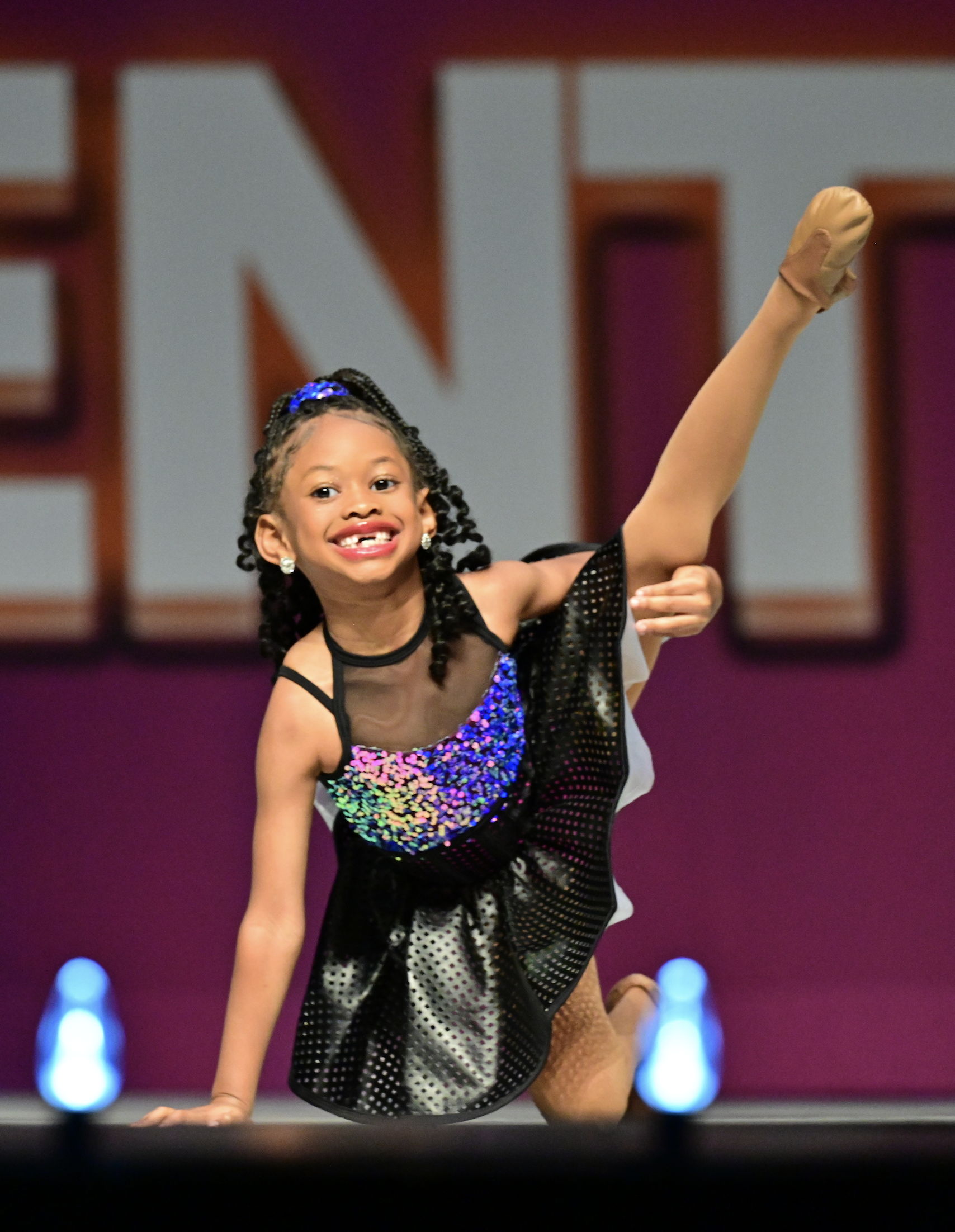 A young girl in a glittery black and multicolored dance costume performs a dance move with her arm raised in front of large purple and black background with the N E T positioned in the background.