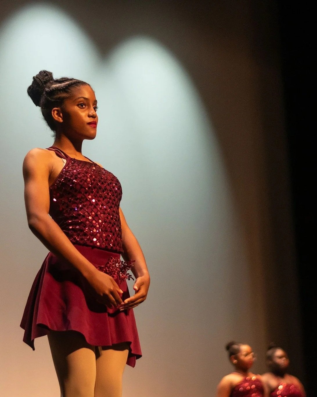 Young girl standing on stage, wearing a sparkling maroon dress with a matching skirt and black tights, illuminated by a spotlight.