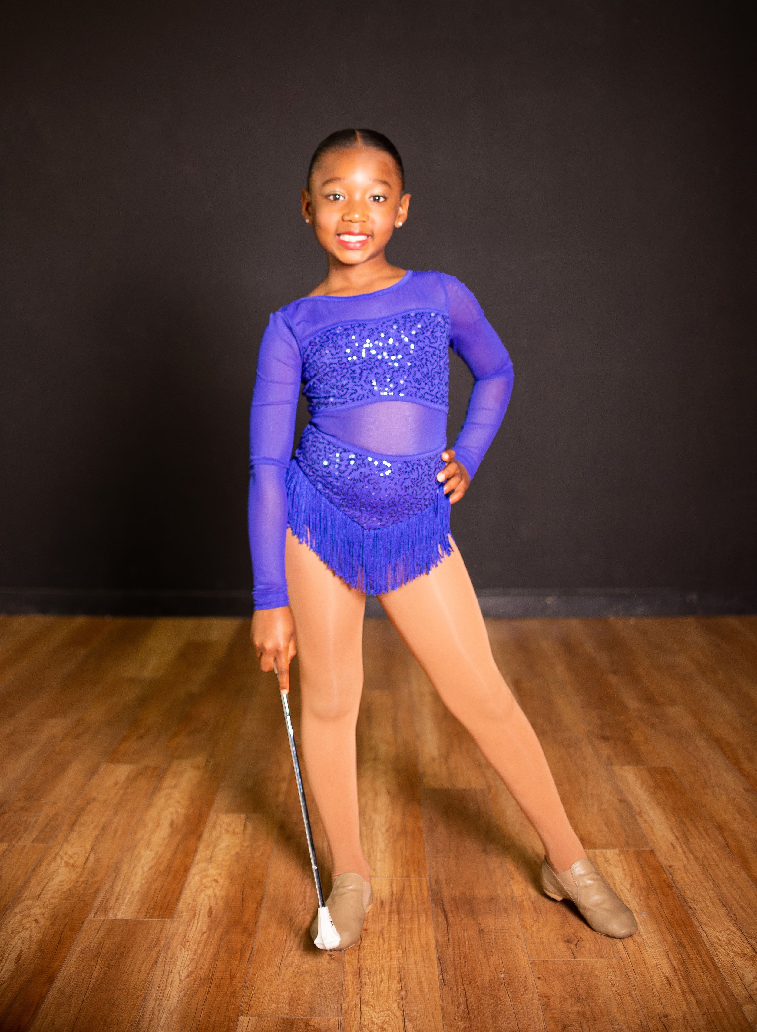 A young girl dressed in a blue rhythmic gymnastics leotard, holding a fencing foil, standing on a wooden floor against a black background, and smiling at the camera.
