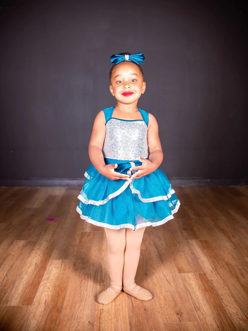 Young girl in a blue and silver dance costume with a bow in her hair, standing on a wooden floor against a dark background.