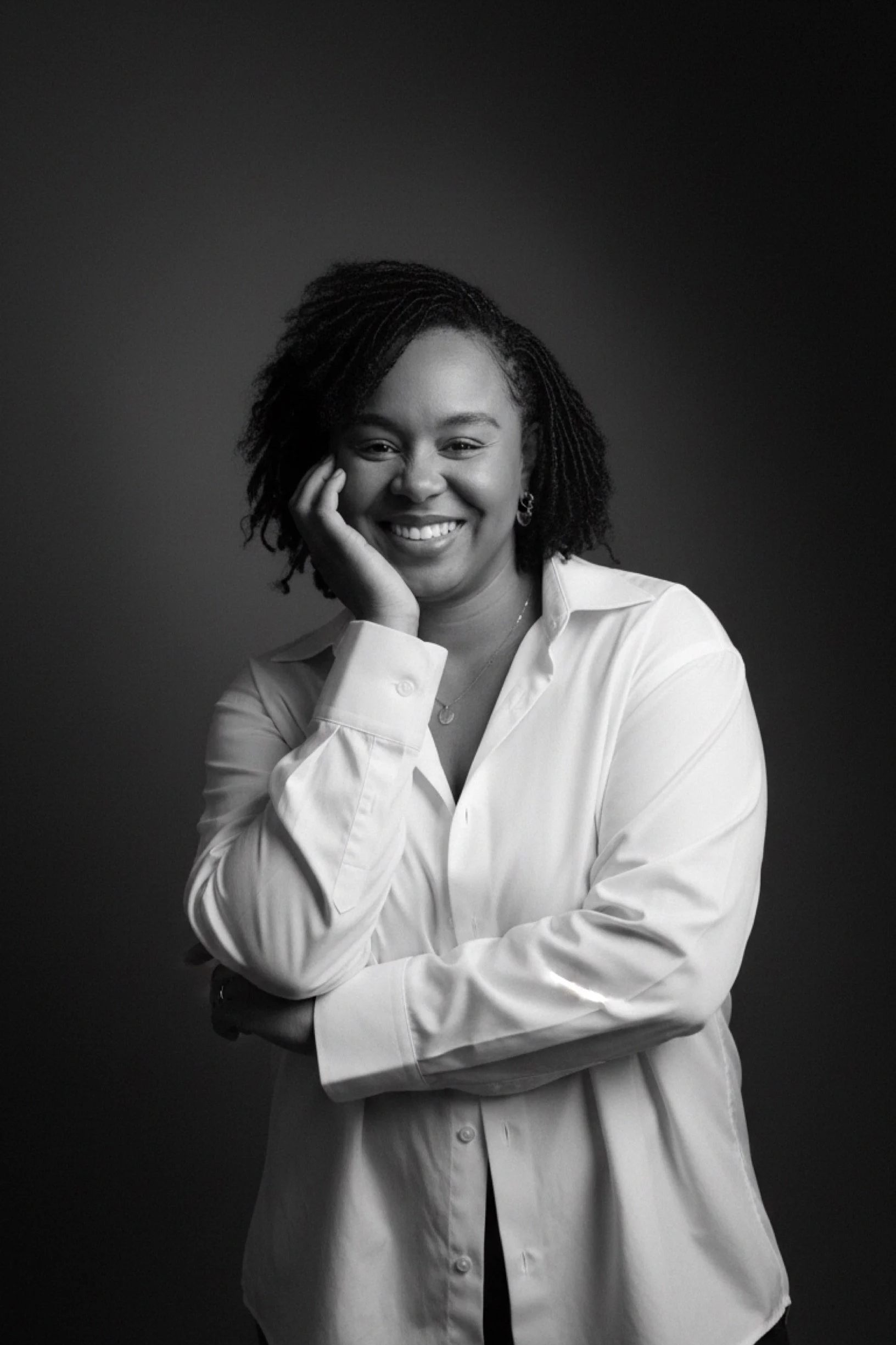 A woman with short, curly hair smiling and posing with her hand on her face, wearing a white button-up shirt, against a dark background.