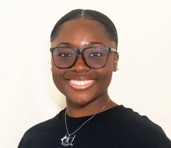 A dance coach in Greensboro, NC smiling African-American woman with glasses, wearing a black shirt and a necklace, against a plain light background.