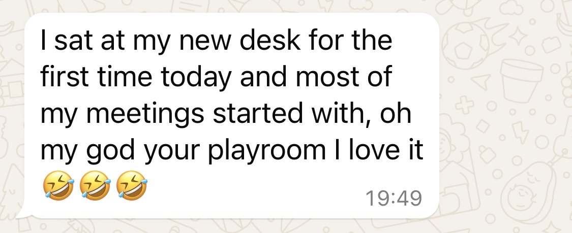 This is a screenshot of a humorous text message about sitting at a new desk for the first time and starting meetings with a comment about a playroom, followed by laughing emojis.