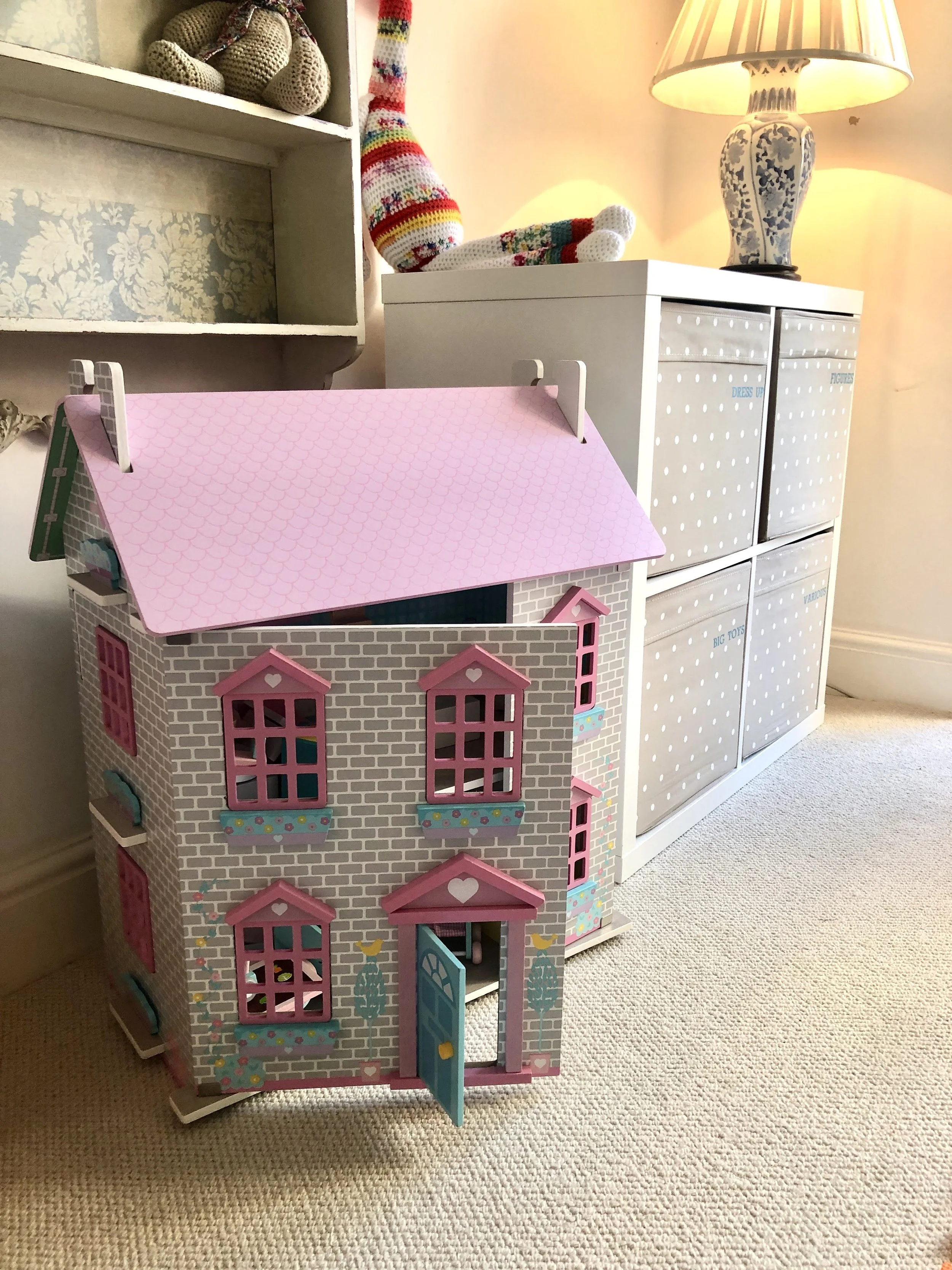 A dollhouse with a pink roof, white brick walls, and pink window frames, placed next to a white storage unit with labeled drawers, decorated with a lamp and stuffed toys on top.