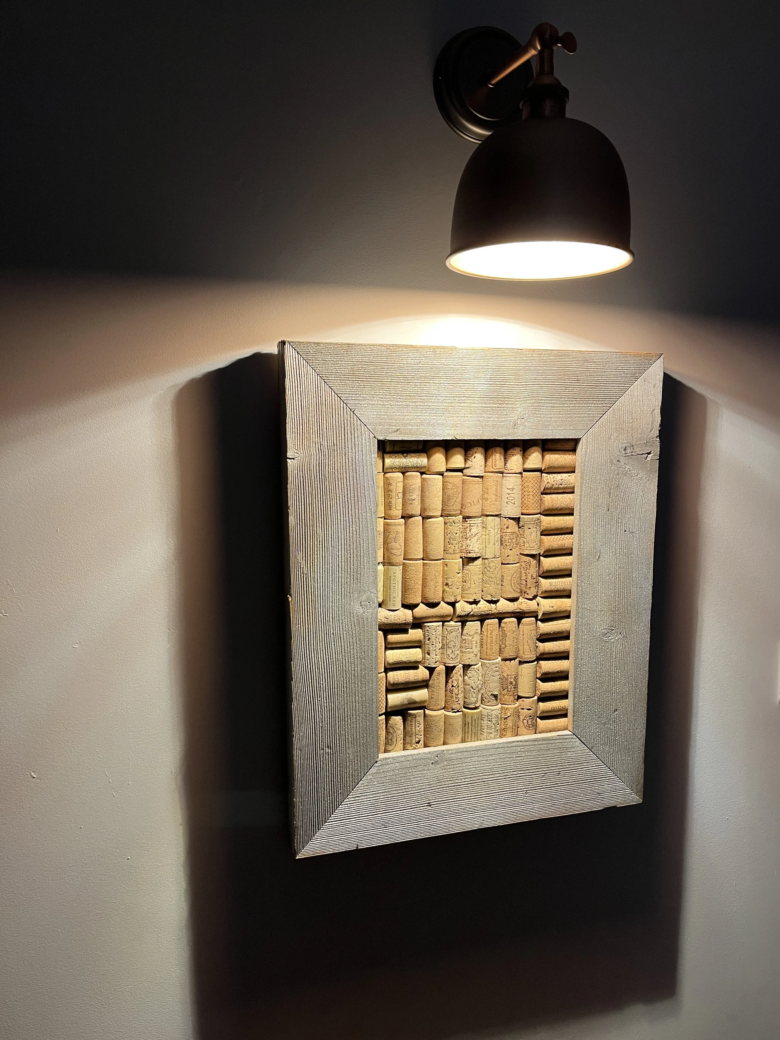 Wall-mounted light shining on a framed collection of wine corks.