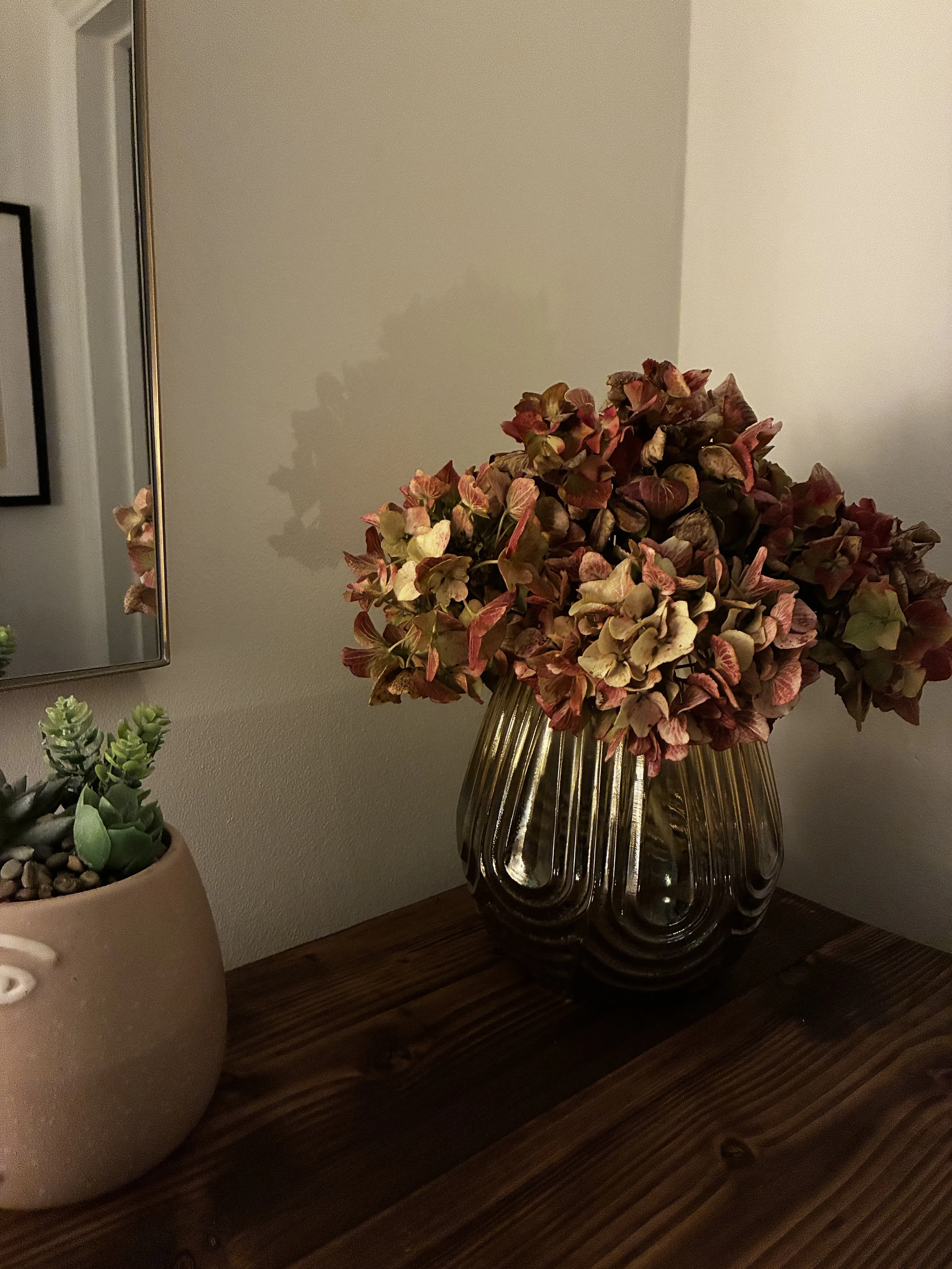 A bouquet of dried hydrangea flowers in a gold-colored glass vase on a wooden surface next to a small potted succulent plant and a mirror on the wall.