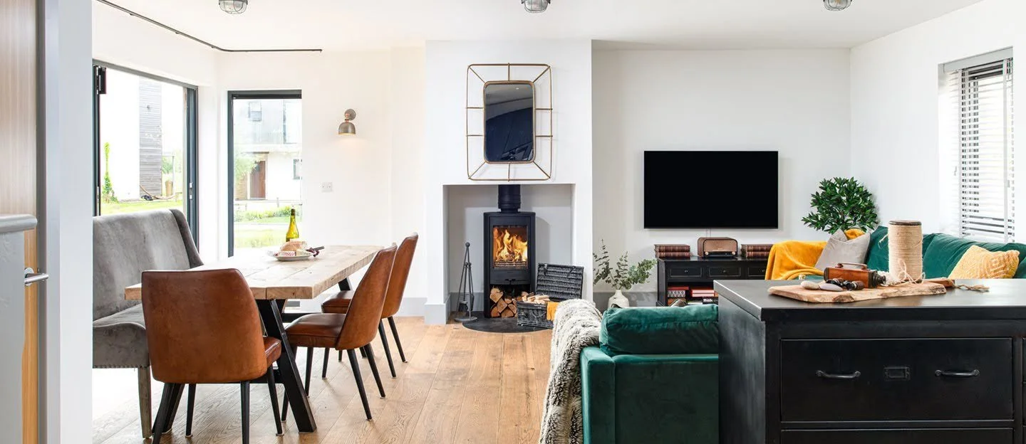 Open-concept living room and dining area with a wood-burning stove, a mounted TV, a large green sofa, and a wooden dining table with mixed chairs, bright natural light from sliding glass doors, and white walls.