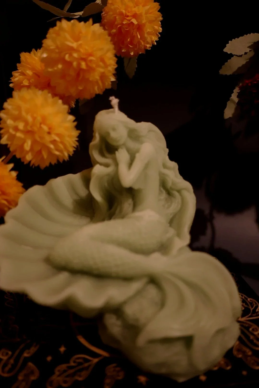A carved candle depicting a mermaid, surrounded by orange chrysanthemum flowers on a dark background.