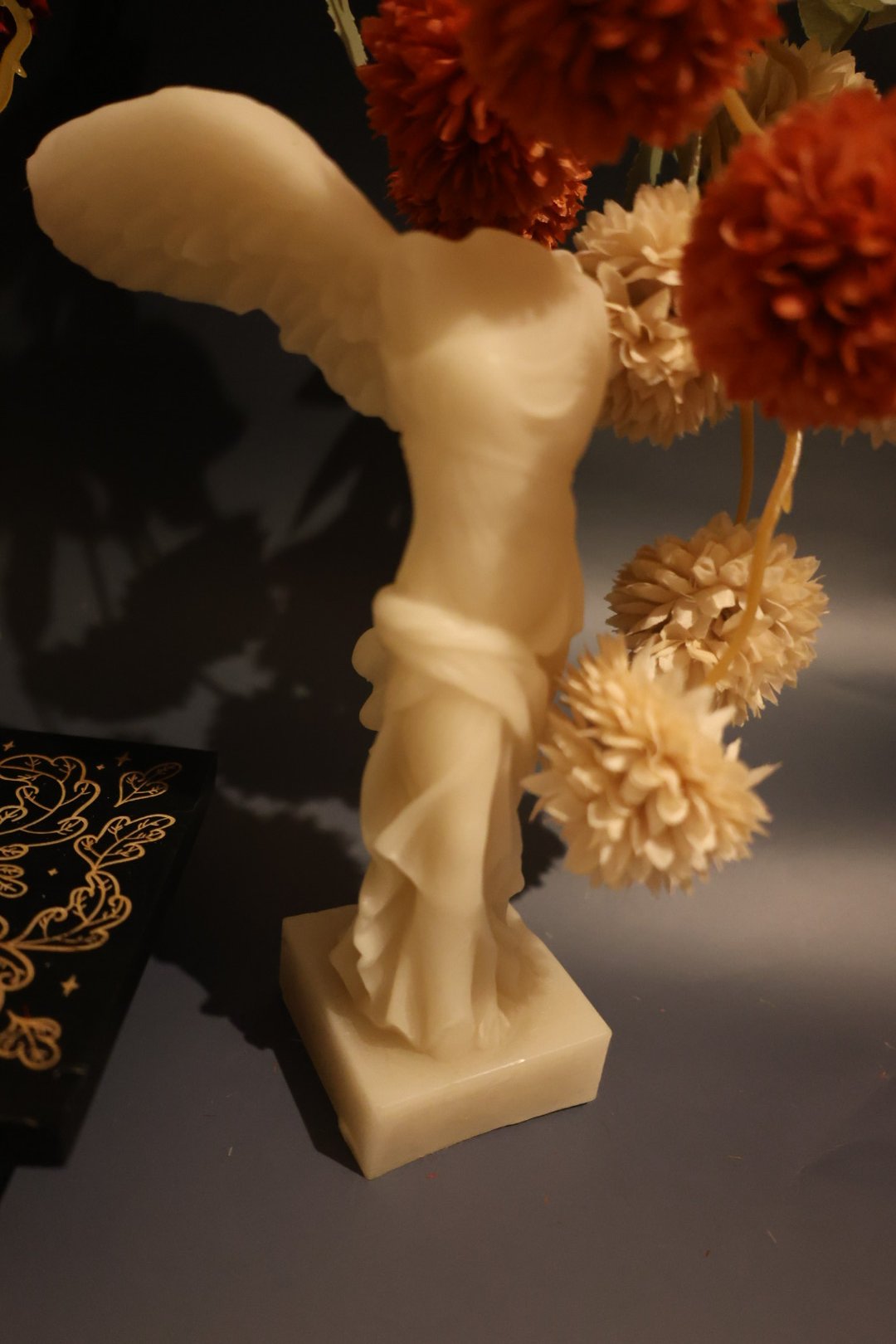 A decorative sculpture of a woman with wings standing on a square base, surrounded by orange and beige artificial flowers.