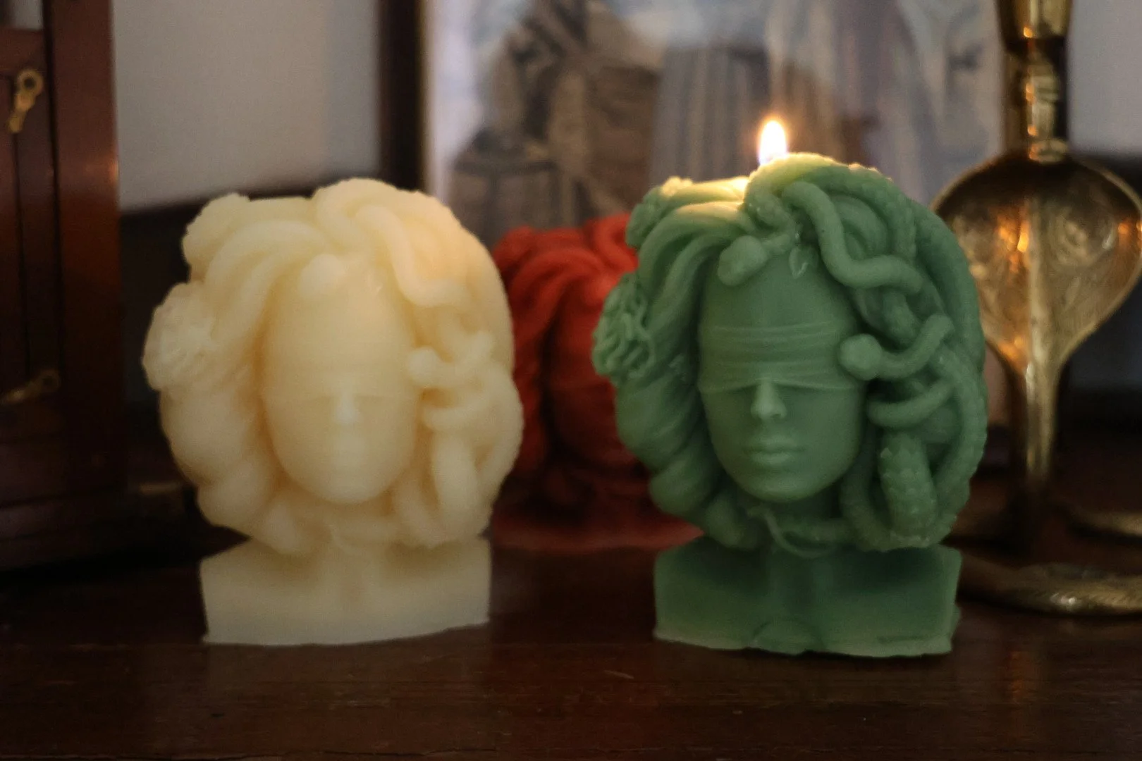 Three decorative candles shaped like human faces with hair, in white, red, and green, placed on a wooden surface with a candleholder and artwork in the background.