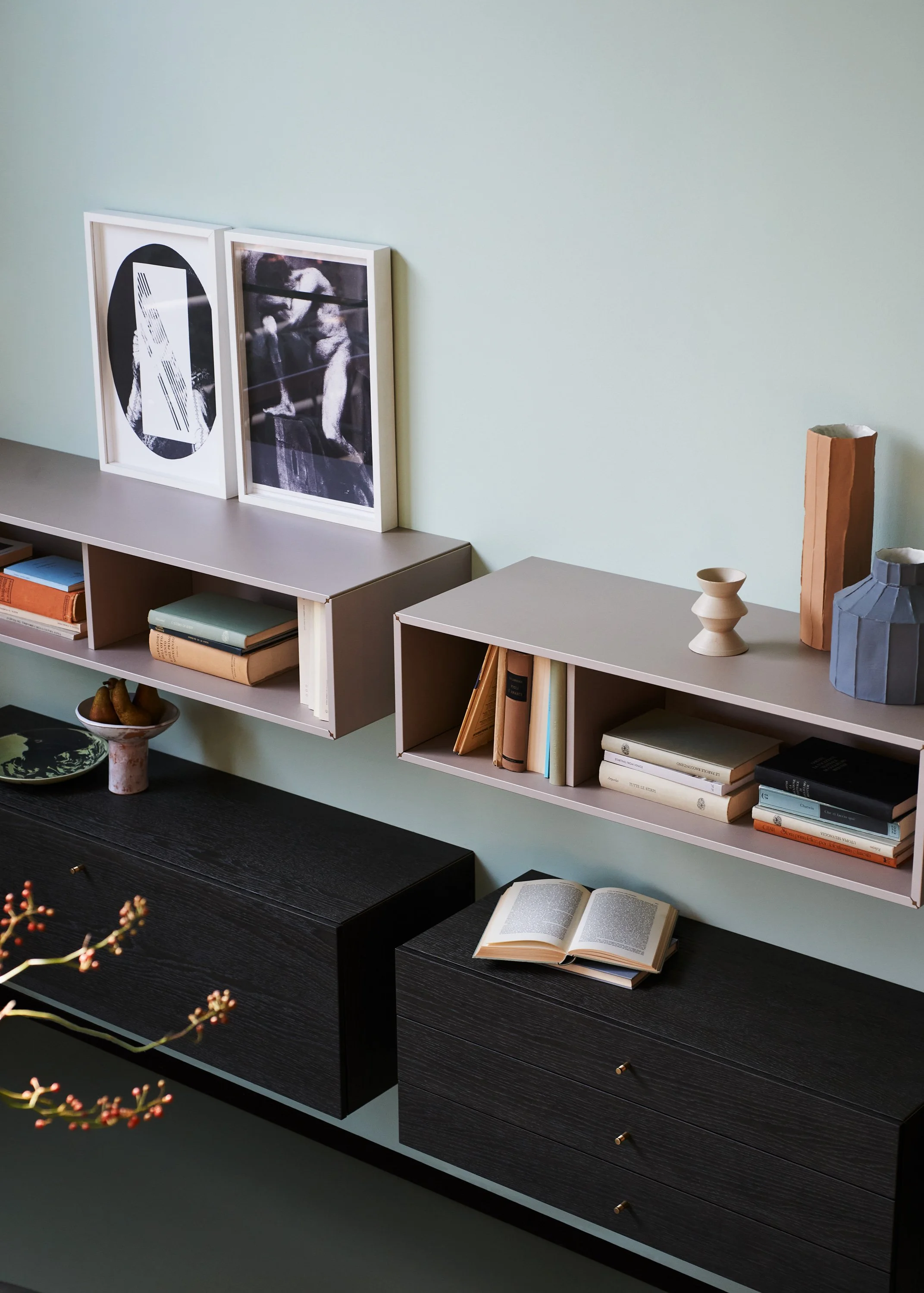 Ein Wohnzimmer mit Wandregalen, deren Oberflächen Bücher, Skulpturen und Kunstdrucke zieren. Es gibt einen schwarzen Couchtisch mit aufgeschlagenem Buch und eine Vase. Im Hintergrund sind Poster in weißen Rahmen. Der Raum hat eine ruhige, moderne Atm