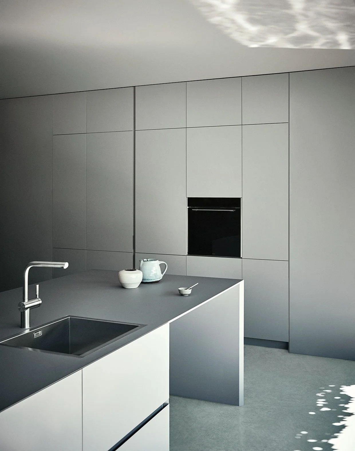 Moderne, minimalistische Küche mit grauen Wänden und Arbeitsflächen, weißen Schränken, Wasserhahn, Teekanne, Schüssel und Löffel auf der Arbeitsfläche, Sonnenlichtmuster auf dem Boden.