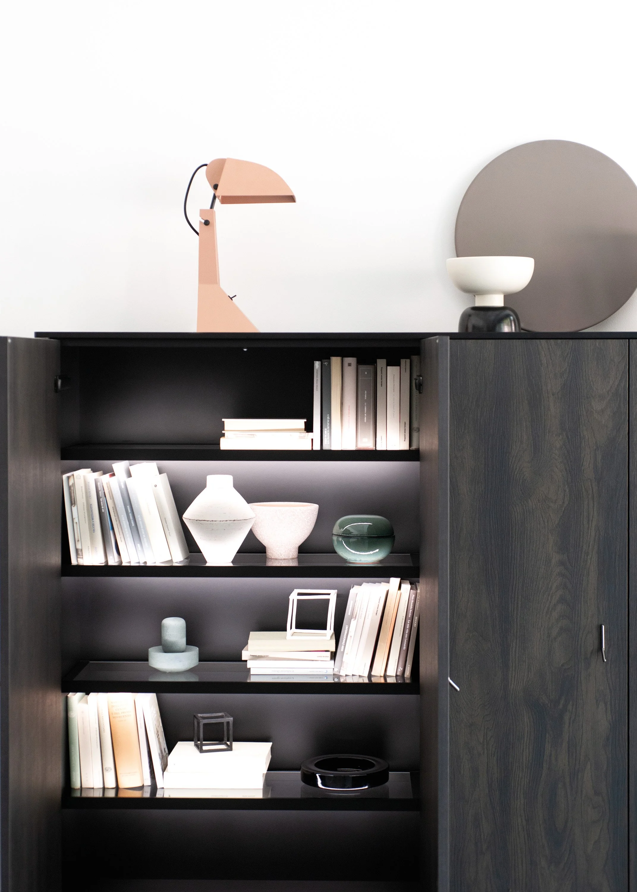 Ein schwarzes Bücherregal mit vier Regalen, die mit Büchern, dekorativen Gegenständen wie Vasen, Rahmen und Skulpturen gefüllt sind. Oben befindet sich ein Tischlampe, darunter eine große runde Dekorationsplatte, sowie Bücher und andere Gegenstände.