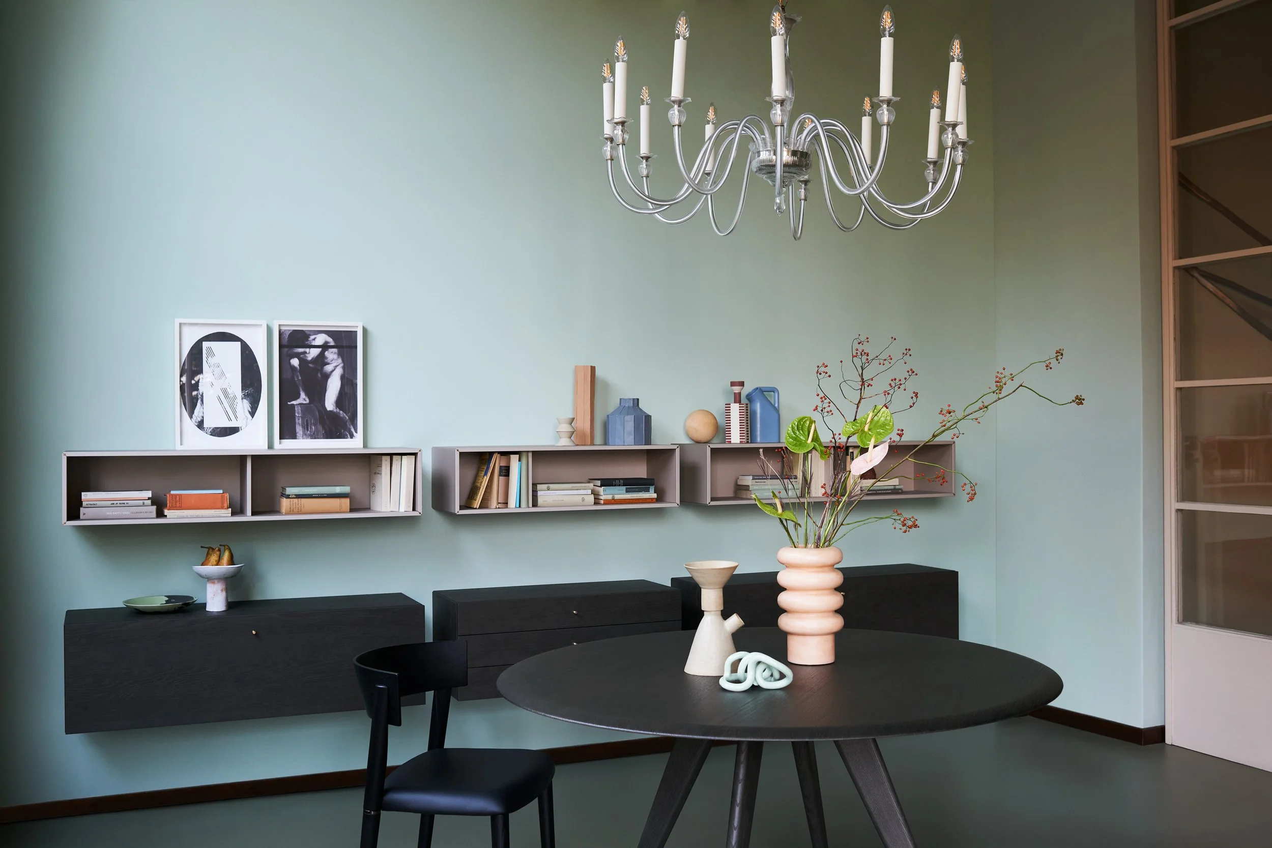 Essbereich mit schwarzem Tisch und schwarzen Stühlen, weißen Wandregalen mit Büchern und Dekoration, moderne Kunstwerke, große Vase mit Zweigen und roten Beeren, grüne Wand in modernem Stil, Schwebeschrank und Deko-Objekte auf dem Regal.