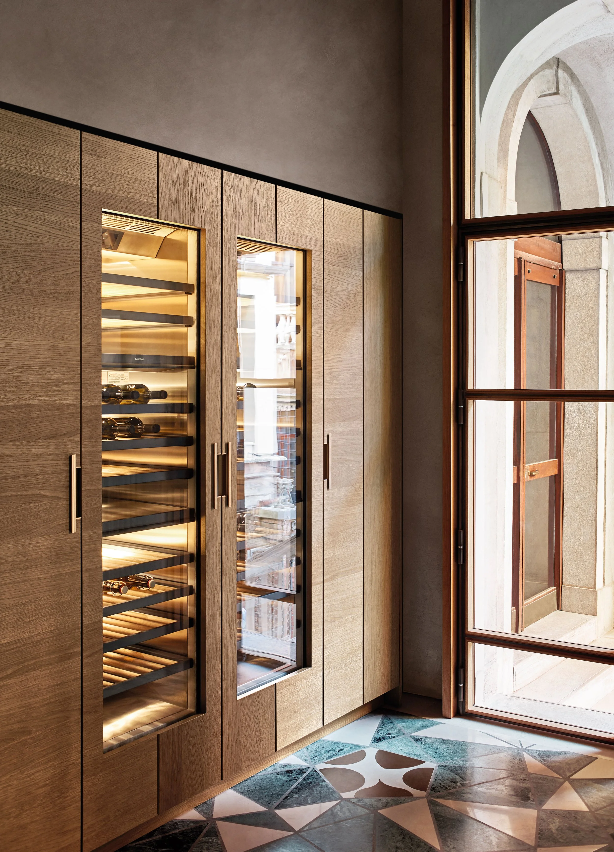 Innenraum mit Holz-Schrank, Weinregal mit Weinflaschen, großer Fenster- und Türaussicht, bunte Fliesenboden.