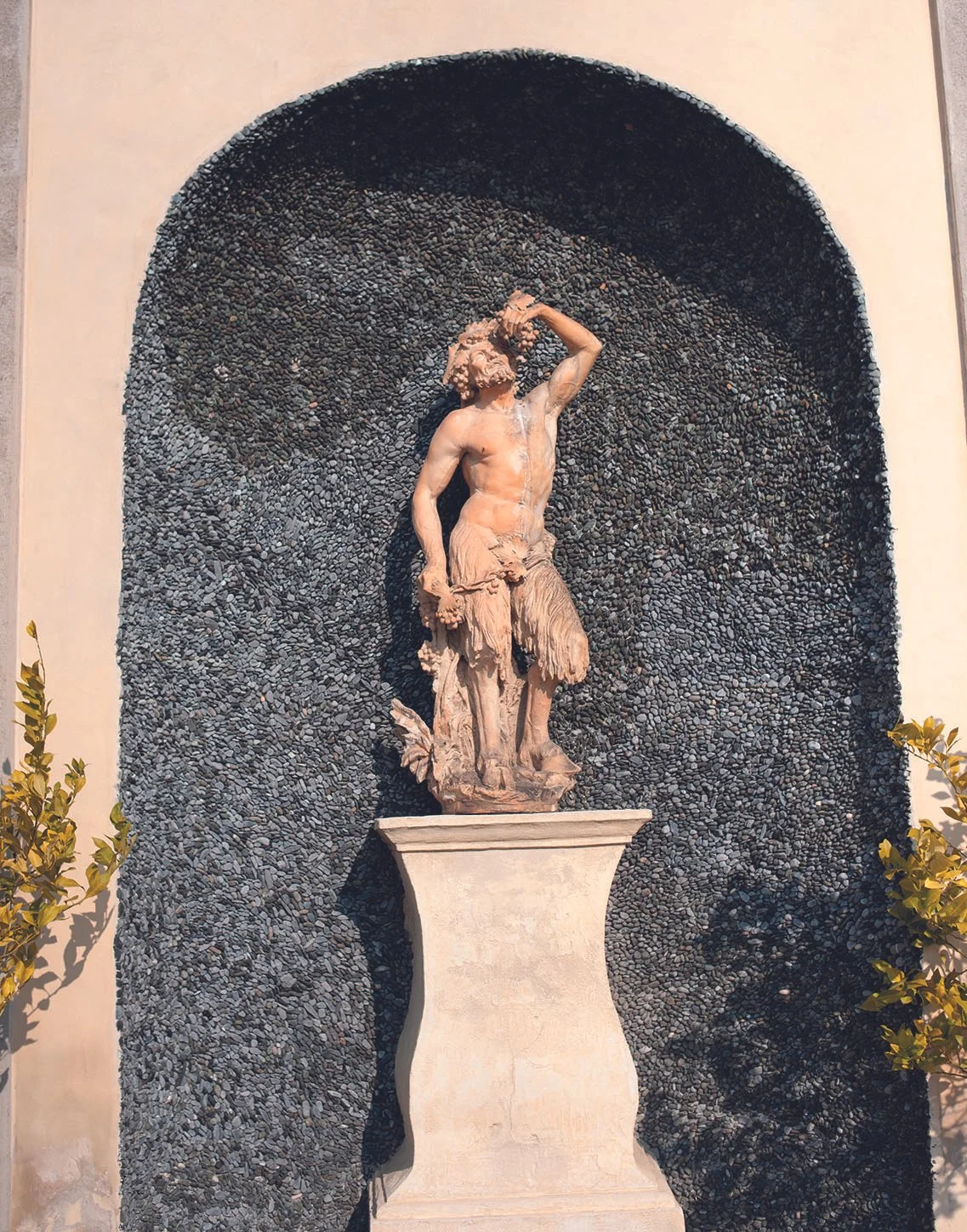 Skulptur eines männlichen Gärtners in einer Nische, umgeben von dunklen Steinen und grünen Pflanzen.