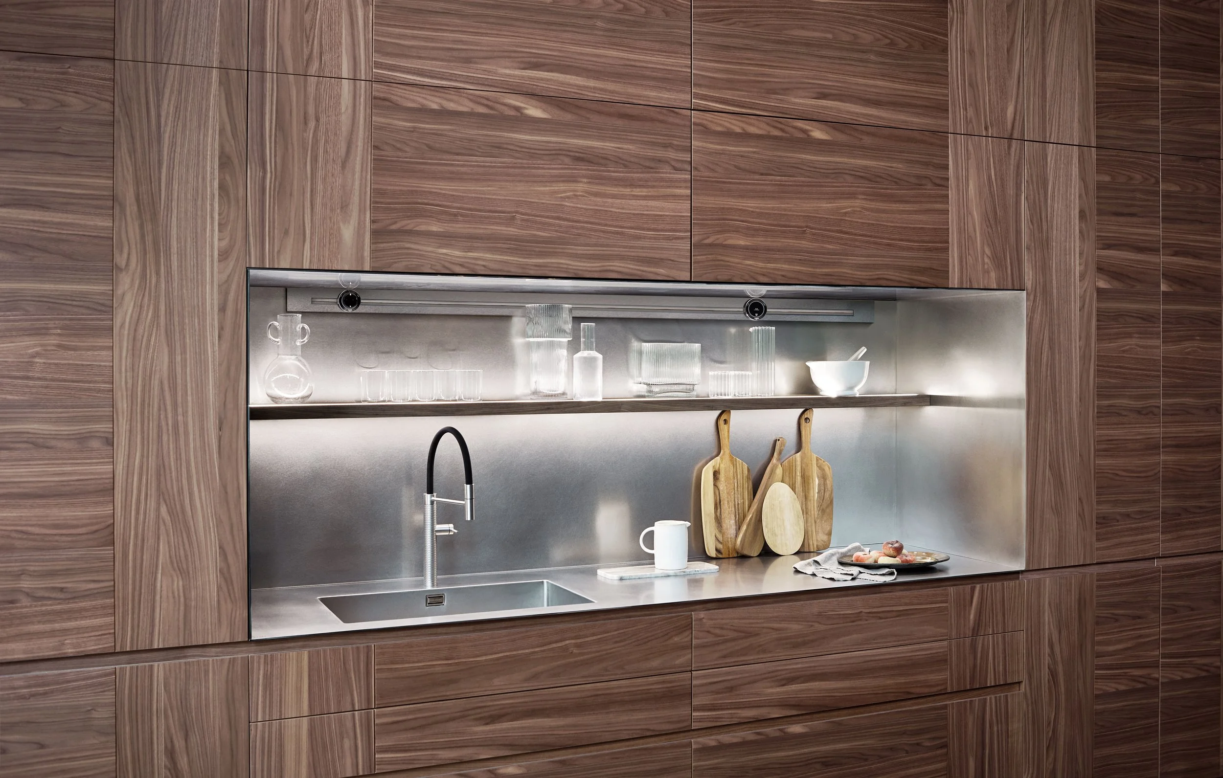 Moderne Küchenschrankwand mit Holzoptik und integriertem Edelstahlspülbecken. Offene Ablage mit Gläsern, Flaschen und Küchenutensilien. Zwei Holzschneidebretter stehen gegen die Wand, daneben eine Tasse und ein Teller mit Speisen.