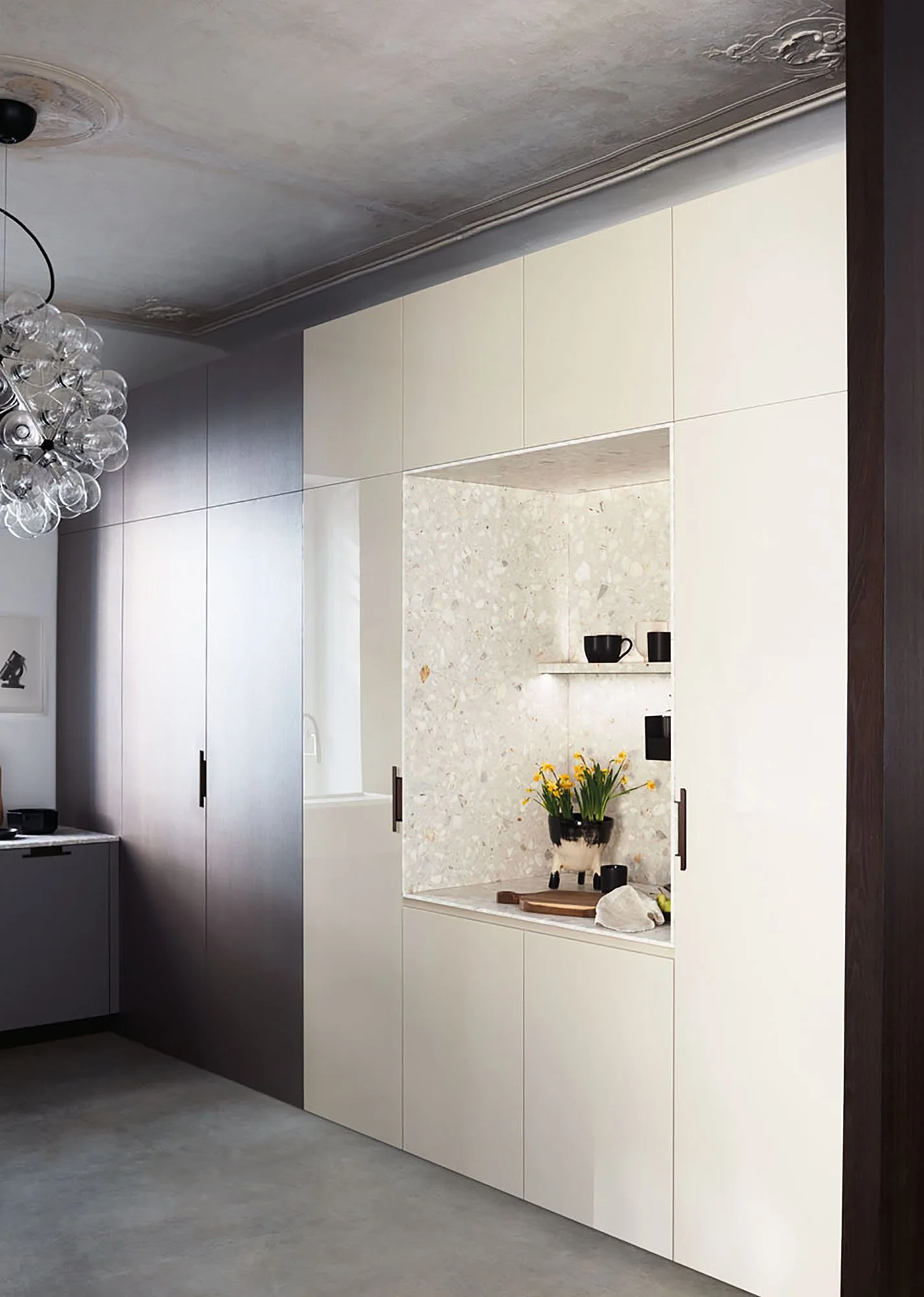 Moderne Küche mit weißen und dunklen Schränken, einer Marmorwandnische mit schwarzen Tassen, einem Blumentopf mit gelben Blumen, einem Holzbrett und Geschirr im Vordergrund, und einer Beleuchtung mit Glühbirnen.