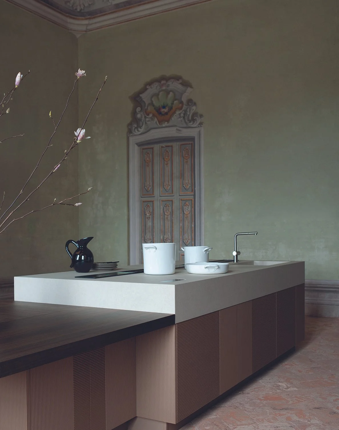 Moderner Küchenbereich mit weißer Insel, schwarzer Kelle, weißen Töpfen und einem Wasserhahn, im Hintergrund eine alte Wand mit dekorativen Elementen und einer Tür, daneben eine Astzweig mit Blüten.