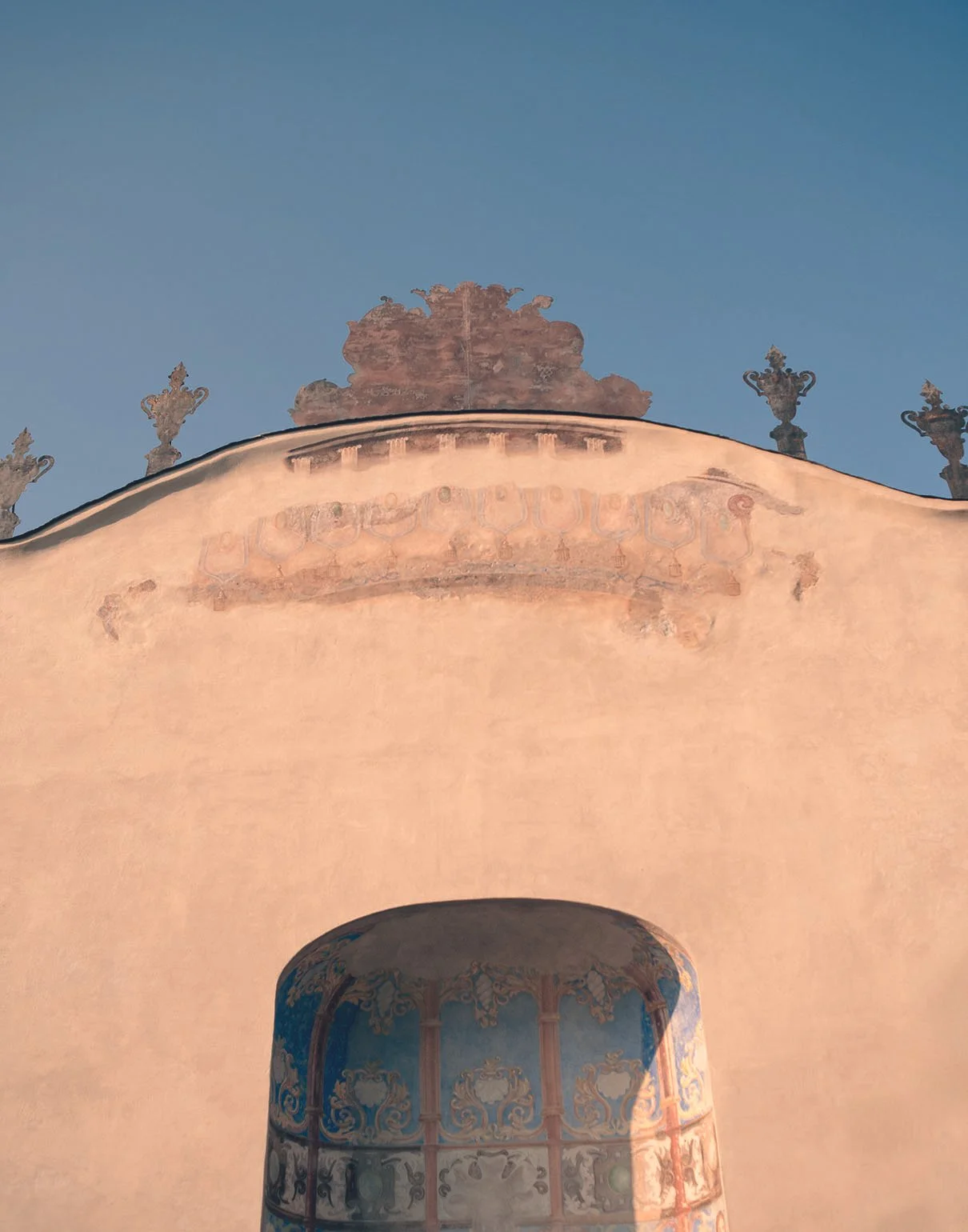 Ein Bauwerk mit einer runden, verzierten Fensteröffnung und dekorativen Ornamenten an der Oberseite, gegen einen blauen Himmel.