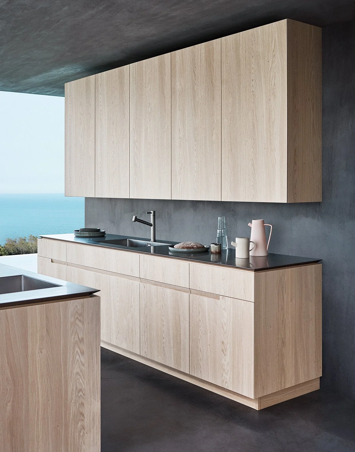 Moderne Kücheneinrichtung mit hellen Holzmöbeln, schwarzer Arbeitsplatte, Wasserhahn, Geschirr, Wasserflasche, Tassen, und Kaffeekanne vor einer Wand mit Blick aufs Meer.