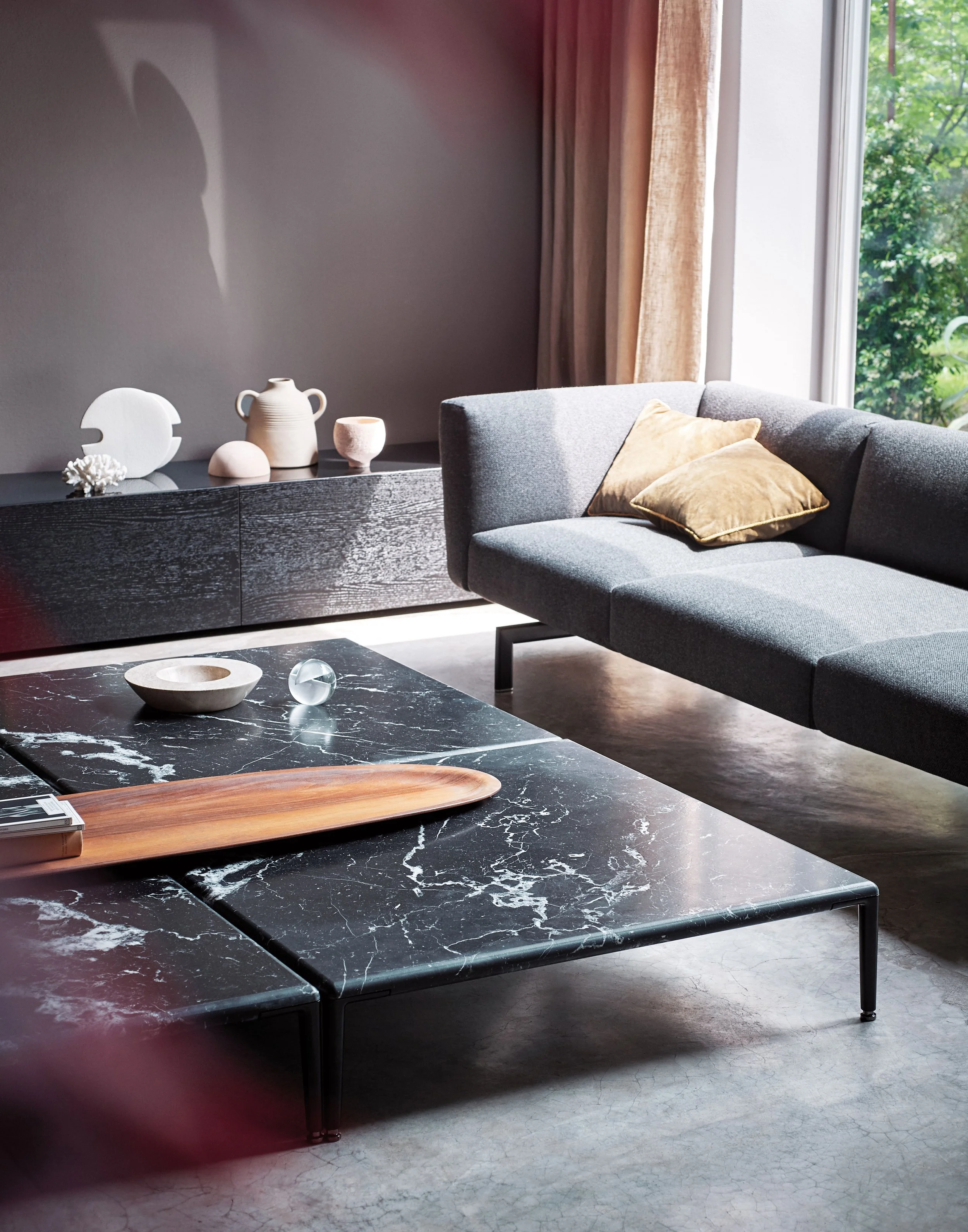 Ein modernes Wohnzimmer mit grauer Couch, beige Kissen, schwarzer Marmortisch und dekorativen Keramiken auf einem schwarzen Sideboard. Große Fenster lassen natürliches Licht herein, draußen sind grüne Bäume sichtbar.