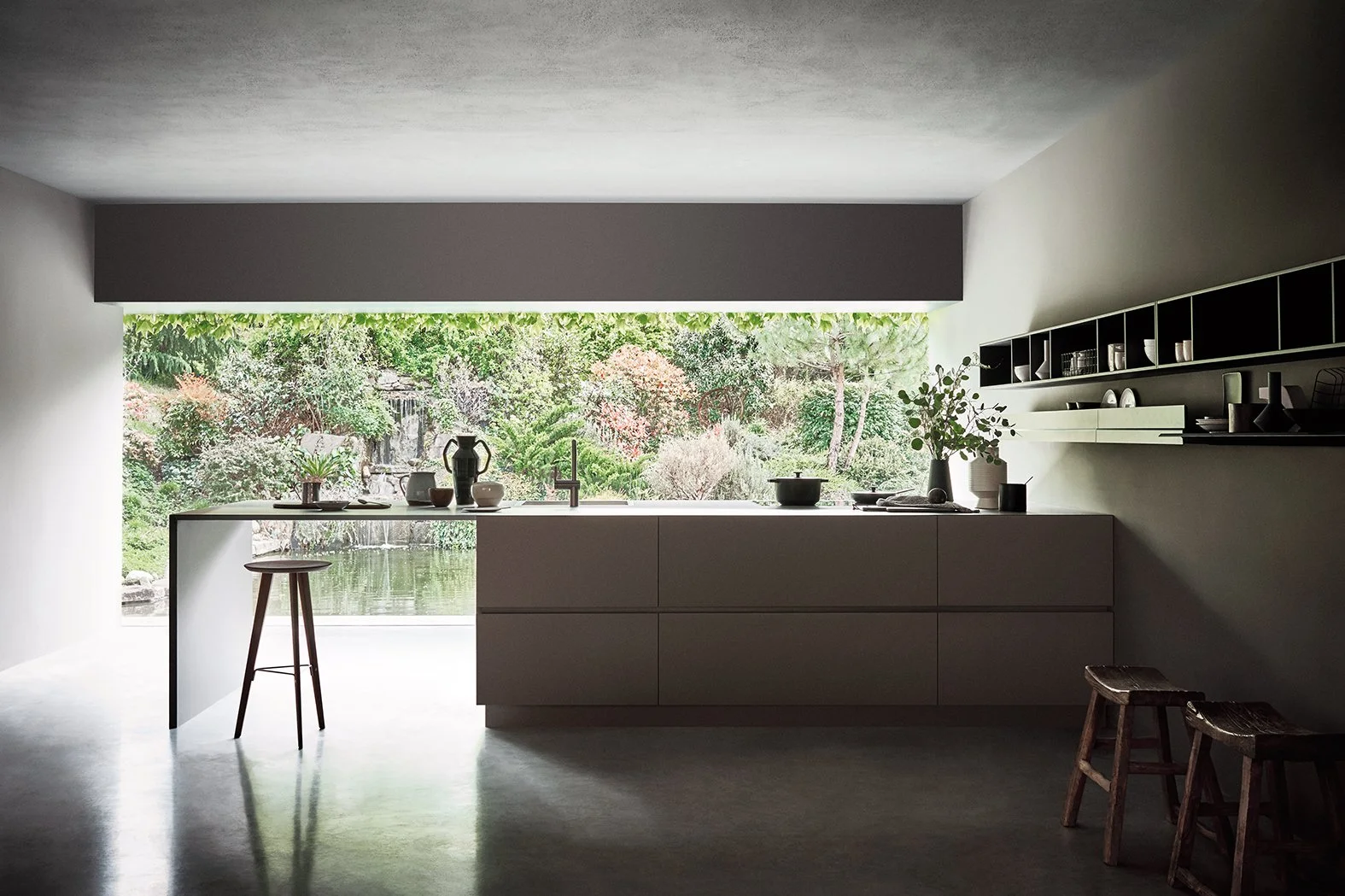 Moderne, minimalistische Küche mit großen Fenster zur Gartenterrasse, mit Küchenutensilien auf der Arbeitsfläche, Pflanzen und Geschirrregale an der Wand.