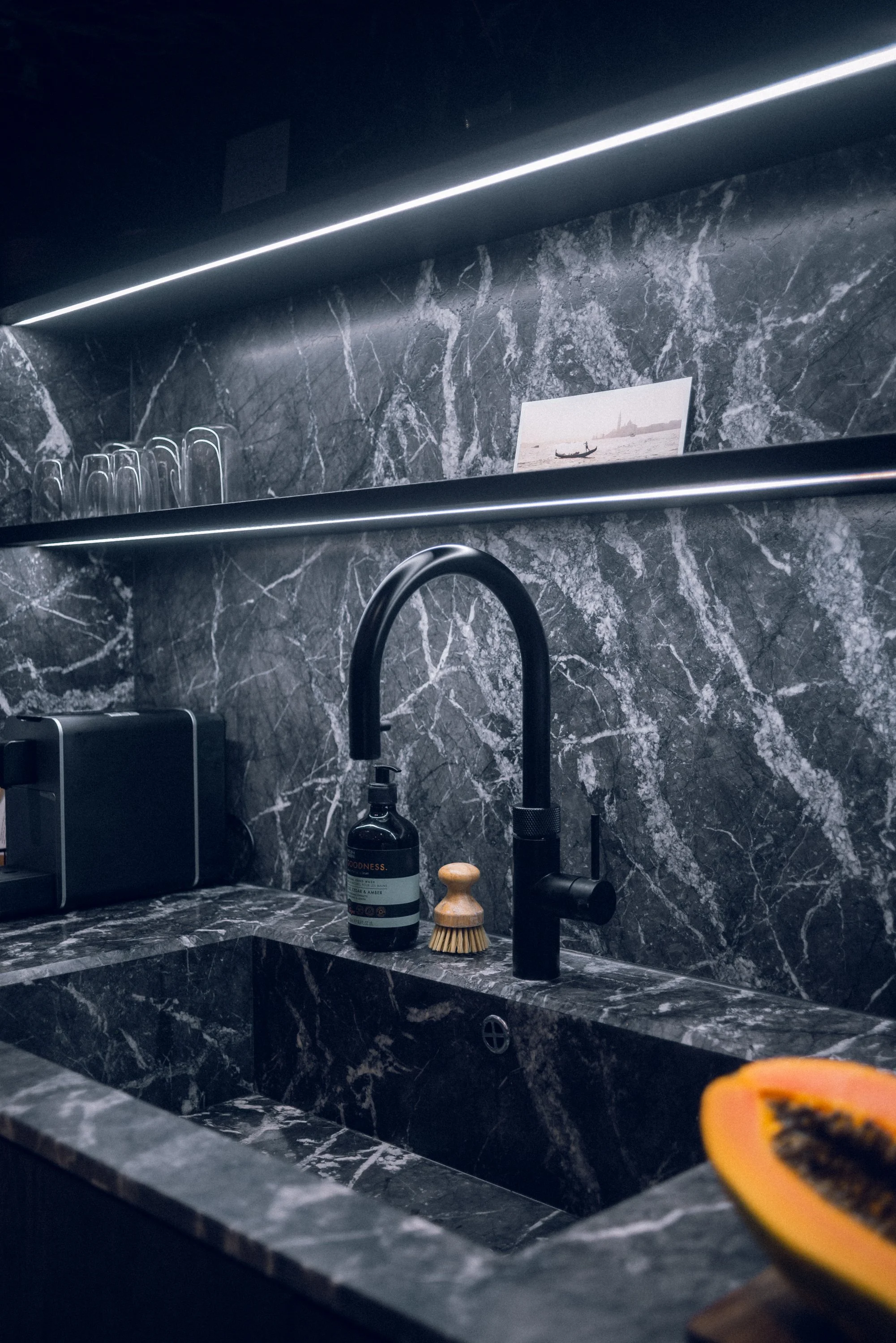 Schwarze Marmor-Küche mit Wasserhahn, Spüle, Geschirrtuch, Papaya auf Schneidebrett, Glasregal mit Gläsern und Dekorationsbild.