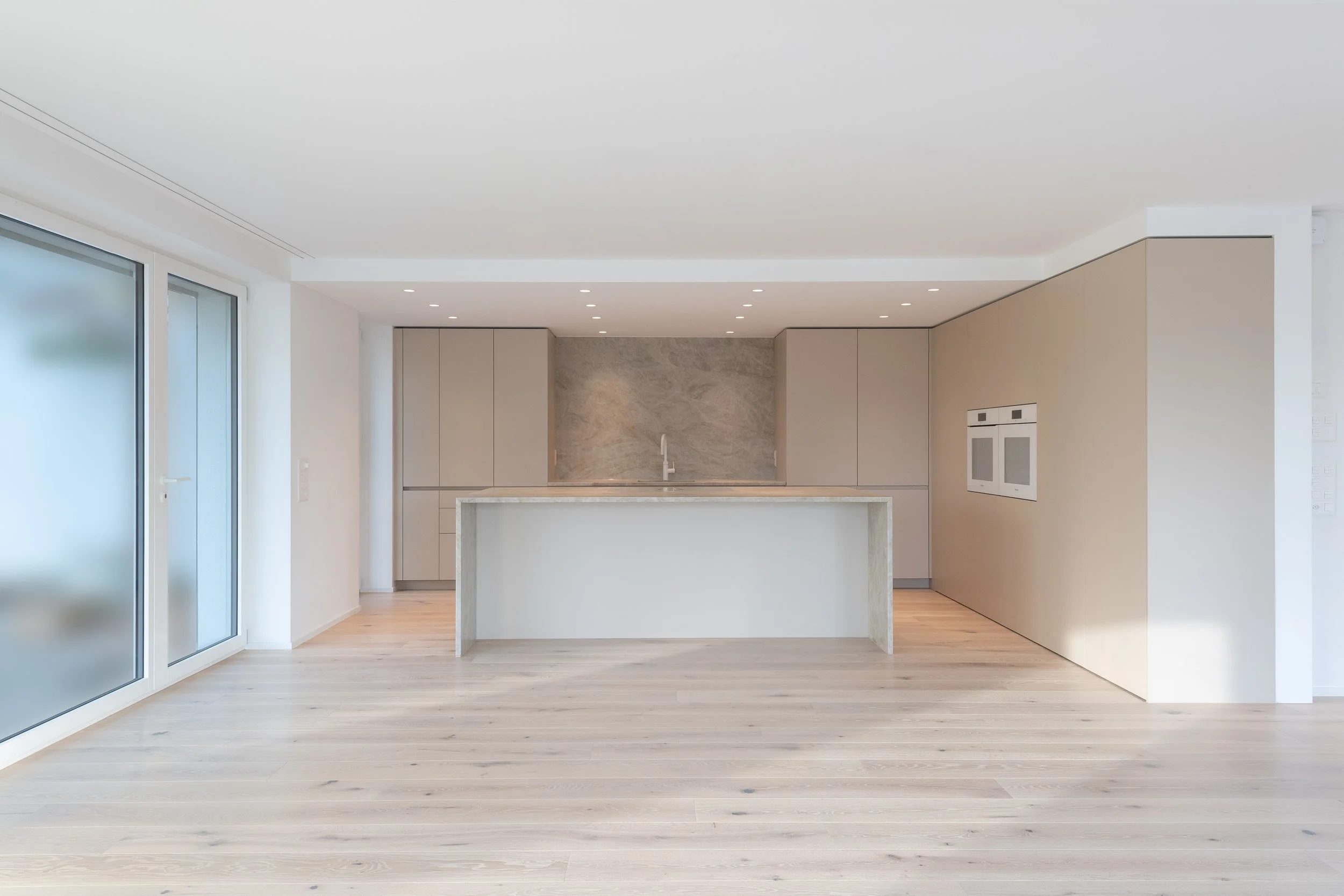Moderne, leere Küche mit weißen und beigen Schränken, Zentralinsel, Marmorwand hinter der Spüle, große Fenster, heller Holzboden, integrierte Geräte.