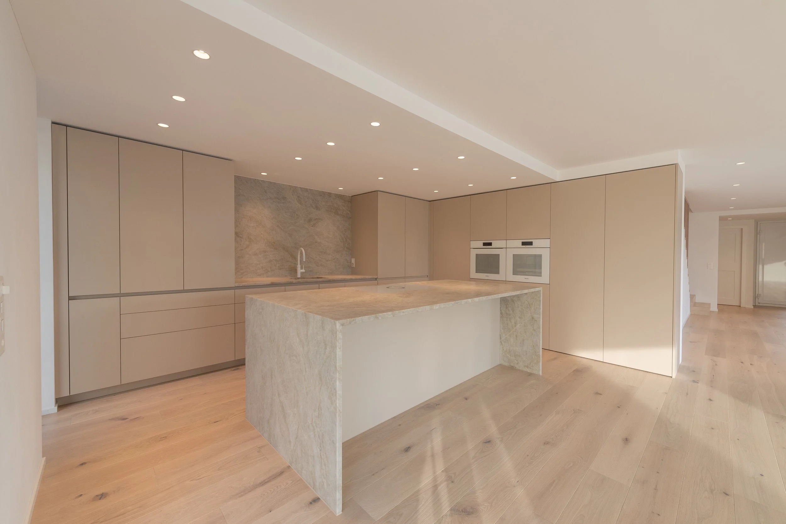 Moderne, helle Küche mit beige Schränken, einer Insel aus hellem Stein, integrierten Backöfen, und einer Holzfußboden.