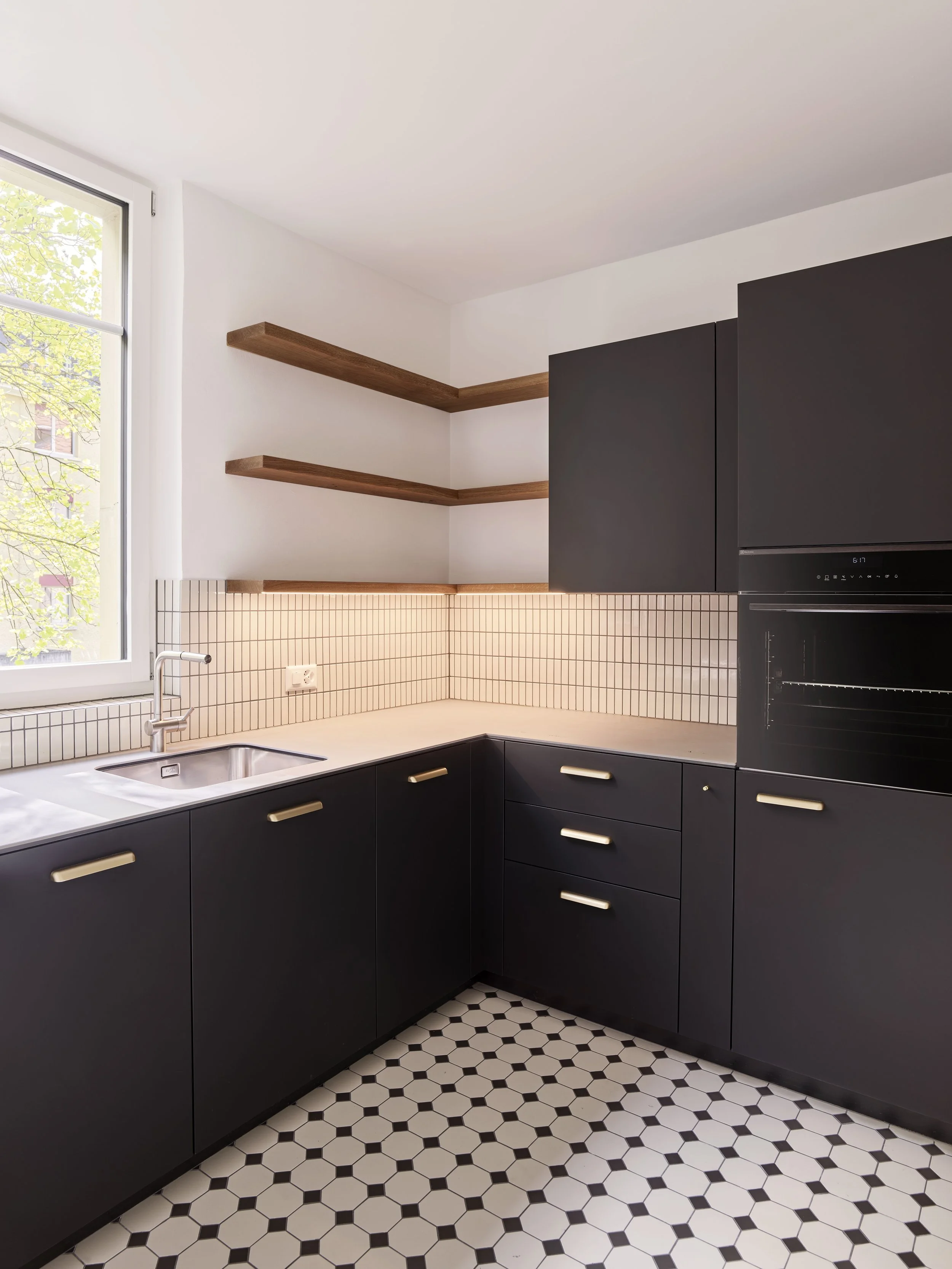 Moderne, minimalistische Küche mit schwarzen Schränken, offenen Holzregalen, weißer Arbeitsfläche, kleiner Spüle, weißem Fliesenspiegel und schwarz-weißen Fliesenboden, großer Fenster