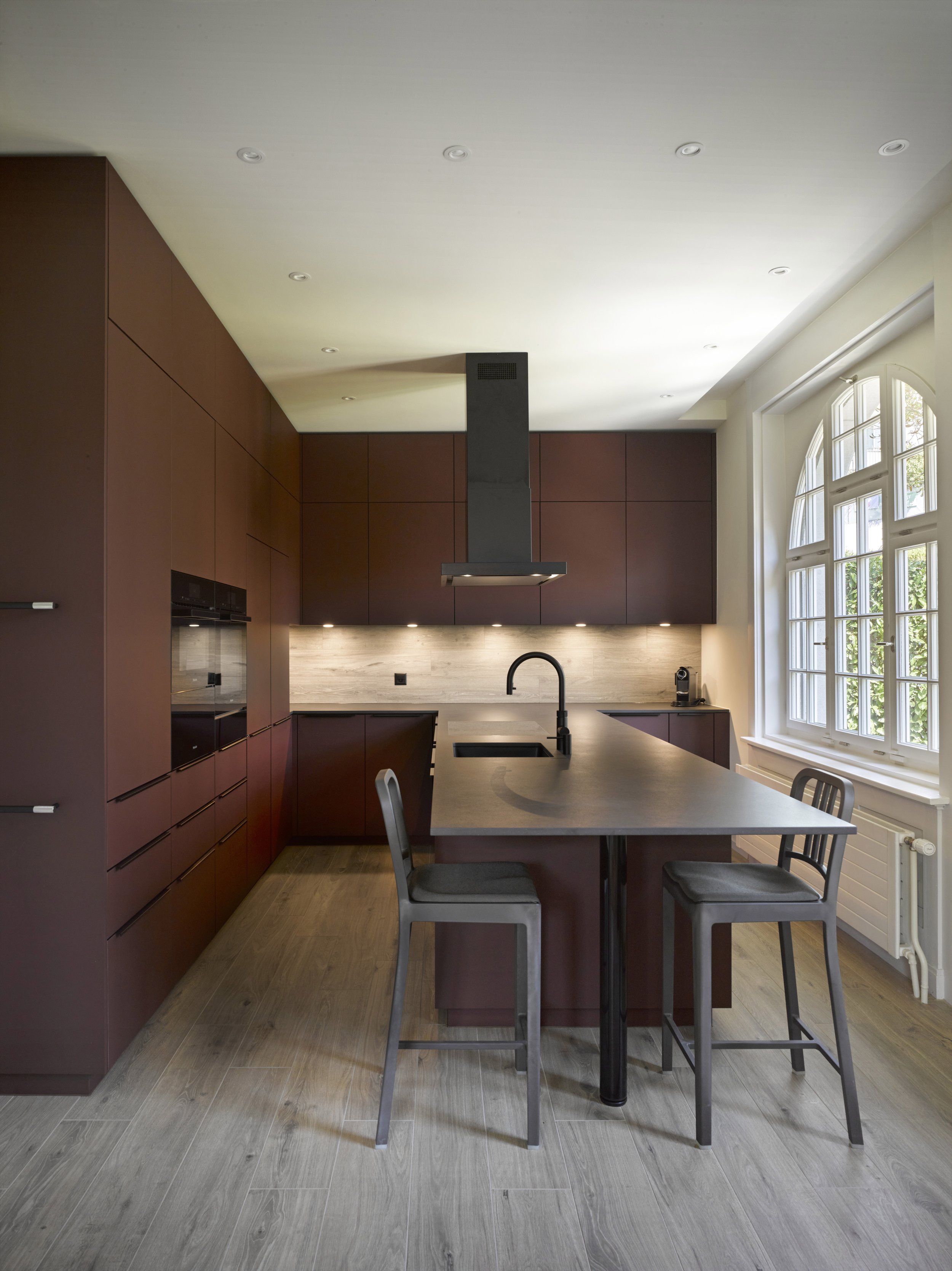 Moderne Küche mit dunklen Schränken, Holzarbeitsplatten, Ofen, Dunstabzugshaube, Wasserhahn, Kaffeemaschine und Sitzgelegenheit am Thekenbereich, große Fenster mit weißen Rahmen
