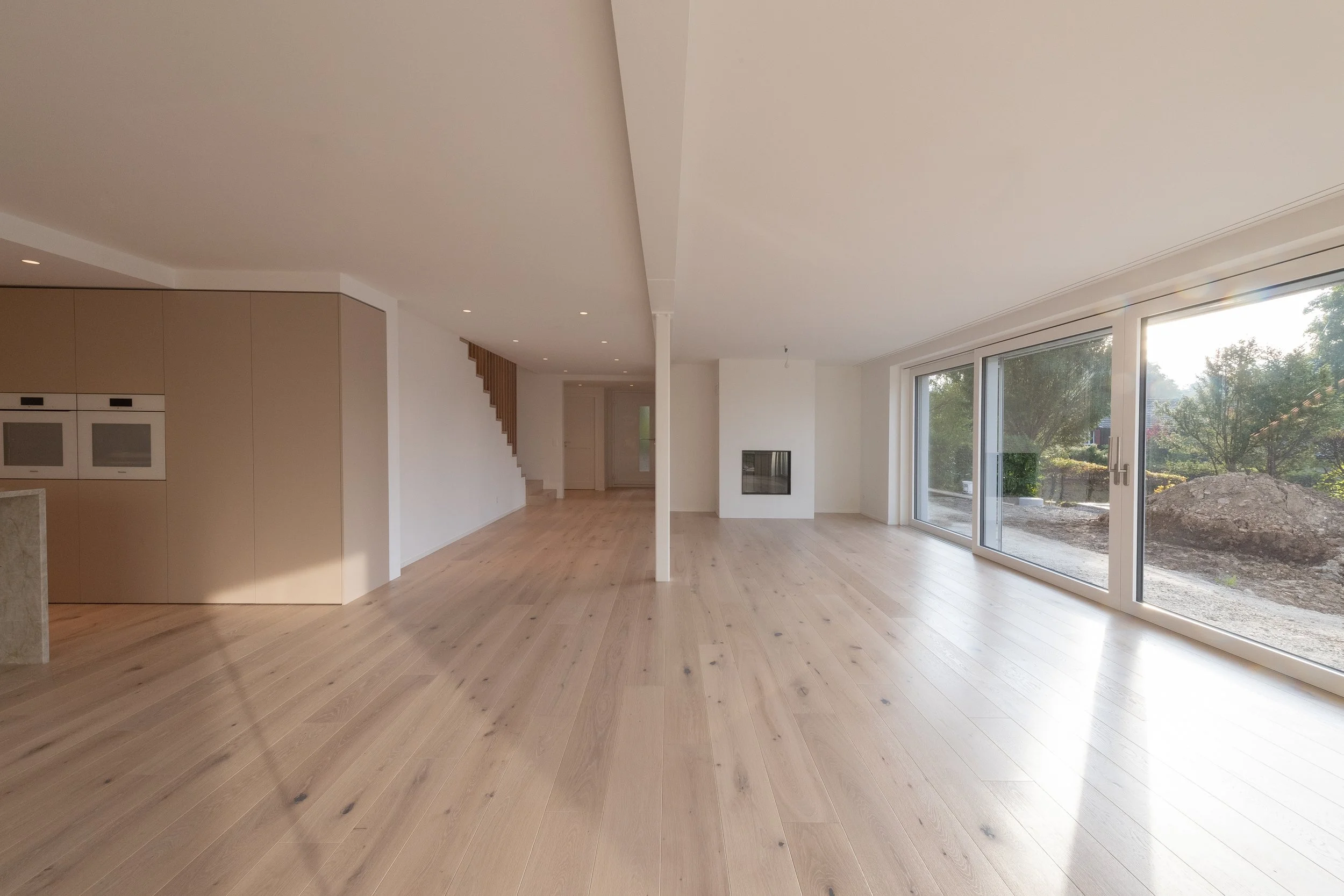 Leeres, modernes Wohnzimmer mit großen Fenstern, Holzboden, Kamin, offener Grundriss, weiße Wände.