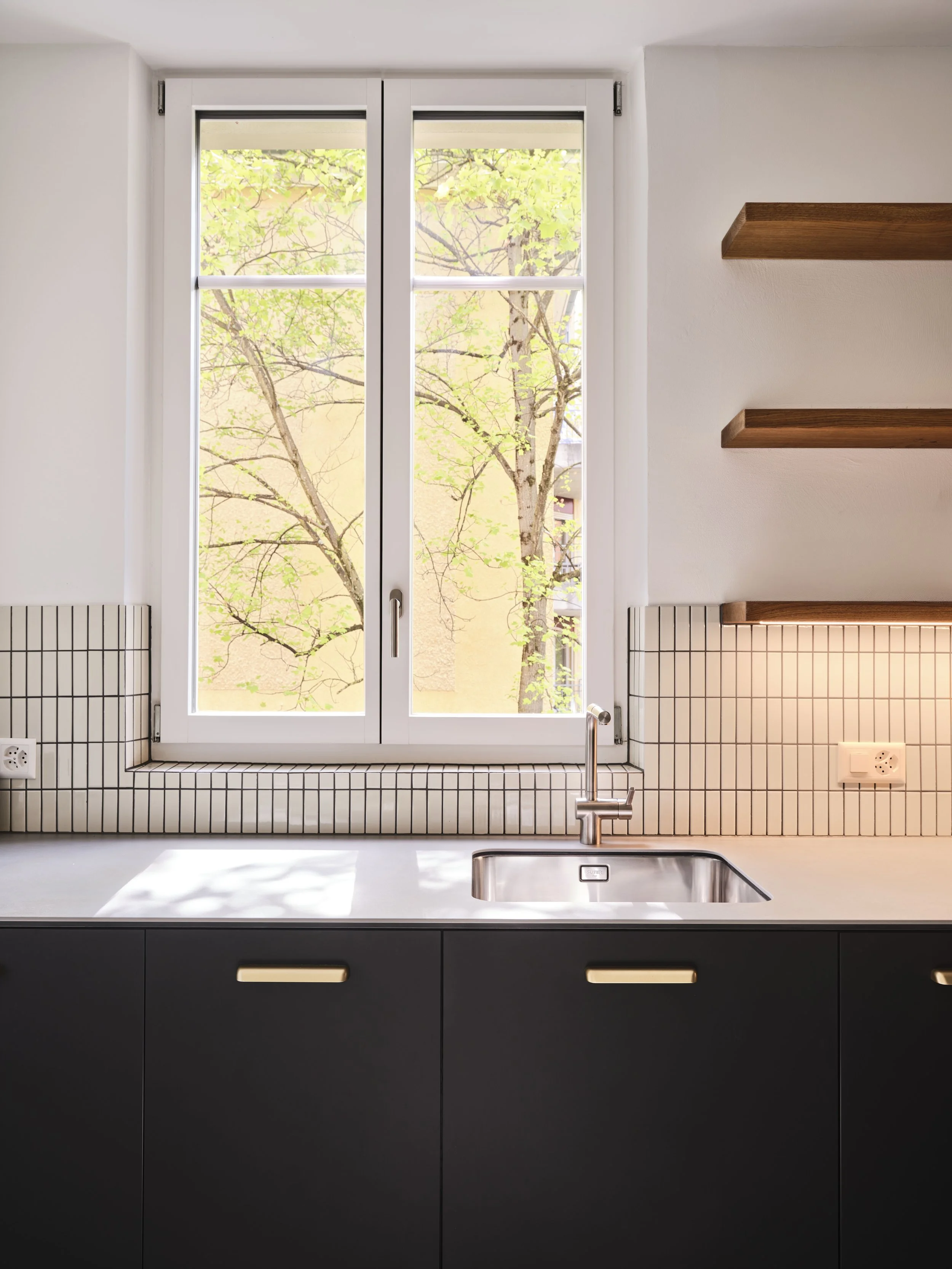 Küchenblick auf ein Fenster mit Baum und grünen Blättern, schwarzer Unterschrank, silberne Spüle, Wasserhahn, weiße Arbeitsplatte, geteilte Steckdosen, Holzregale an der weißen Wand.