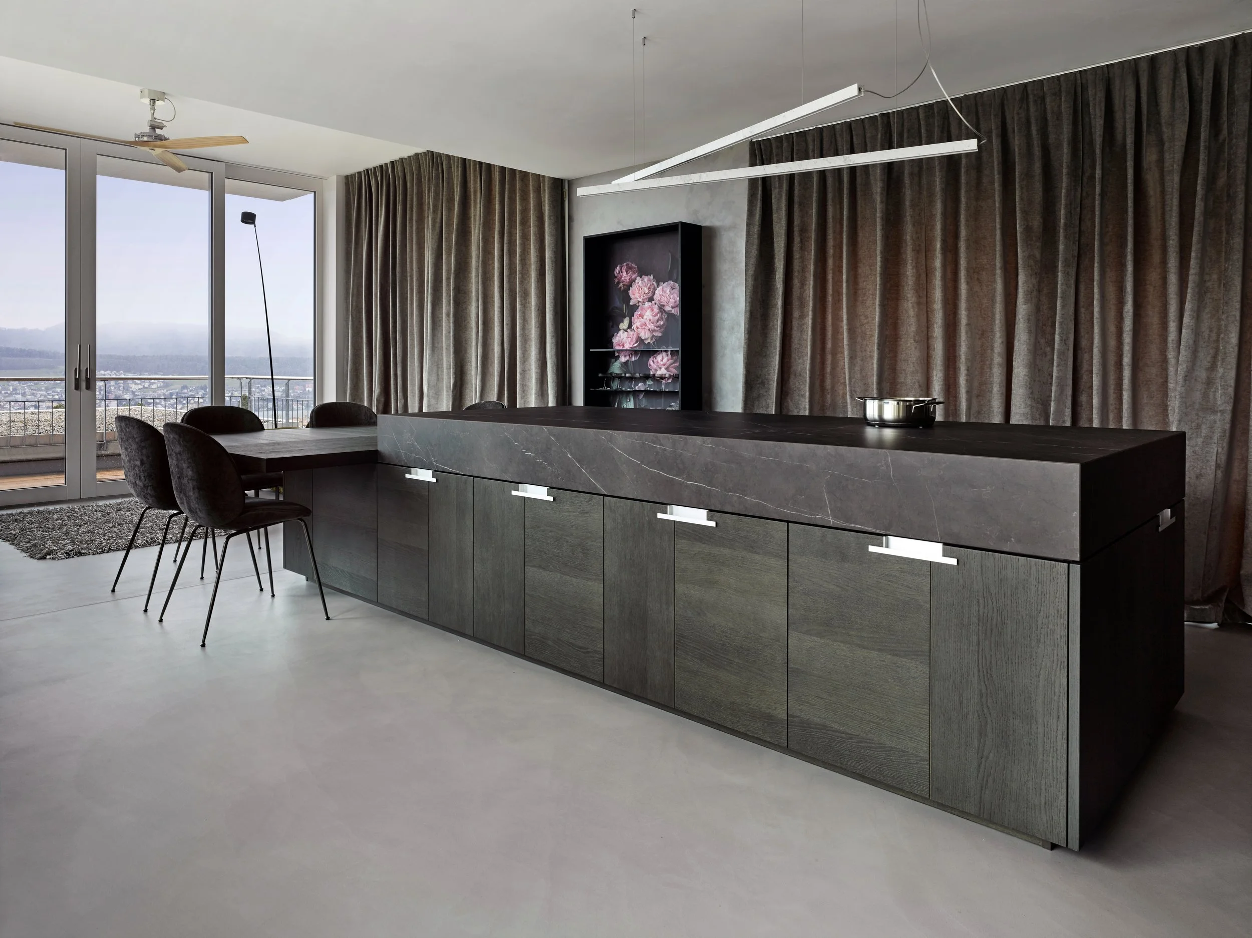 Moderne Küche mit dunklem Holz- und Stein-Design, große Fenster mit Blick auf eine Stadt, lange Küchenzeile mit Herd, schwarze Barhocker, graue Vorhänge, abstrakte Wandkunst, minimalistische Beleuchtung.