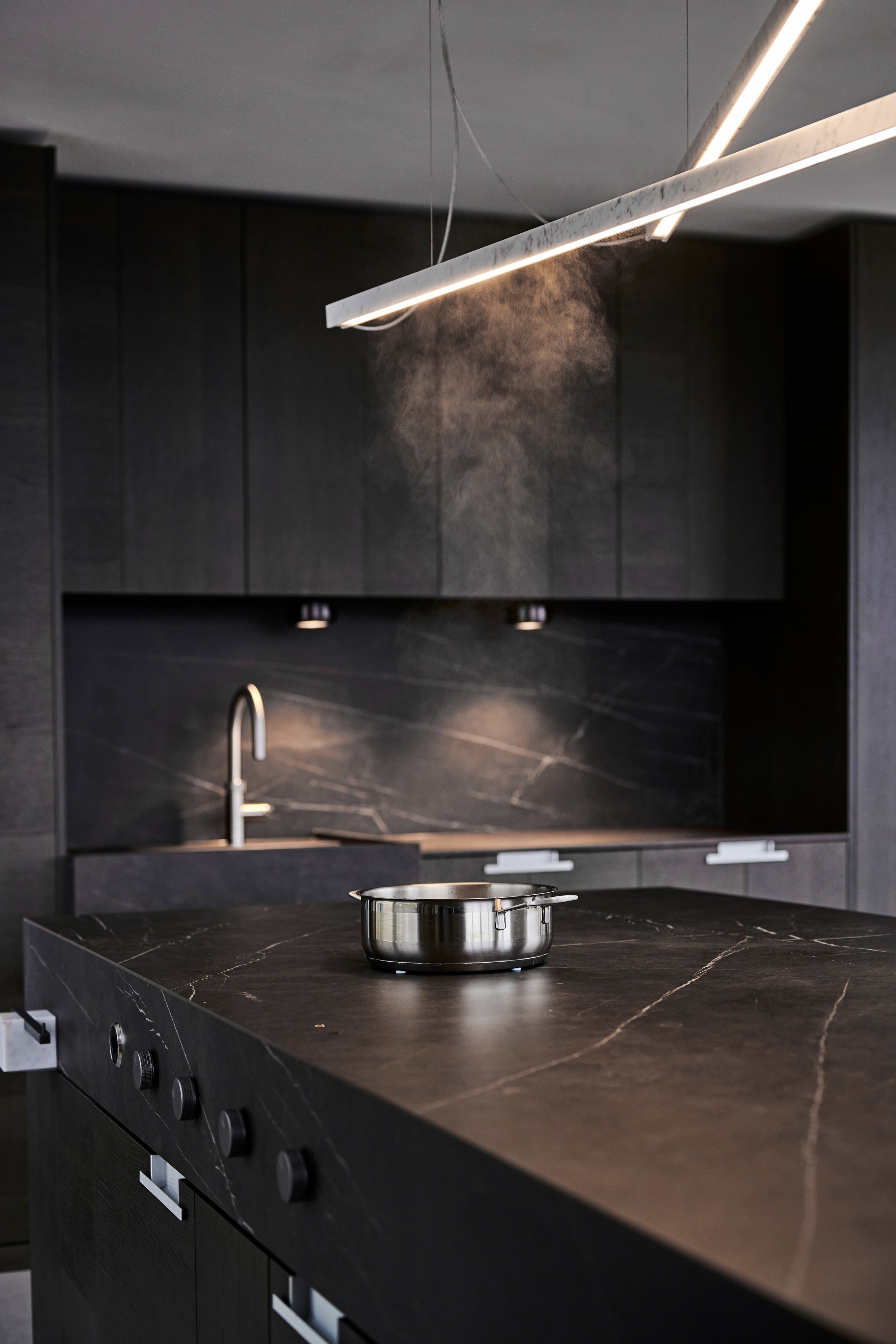 Moderne schwarze Küche mit schwarzen Marmorarbeitsplatten, schwarzem Schrank, Edelstahl-Topf auf der Arbeitsfläche, undermontierter Wasserhahn und hängender Leuchte, die Staub oder Dampf in der Luft sichtbar macht.