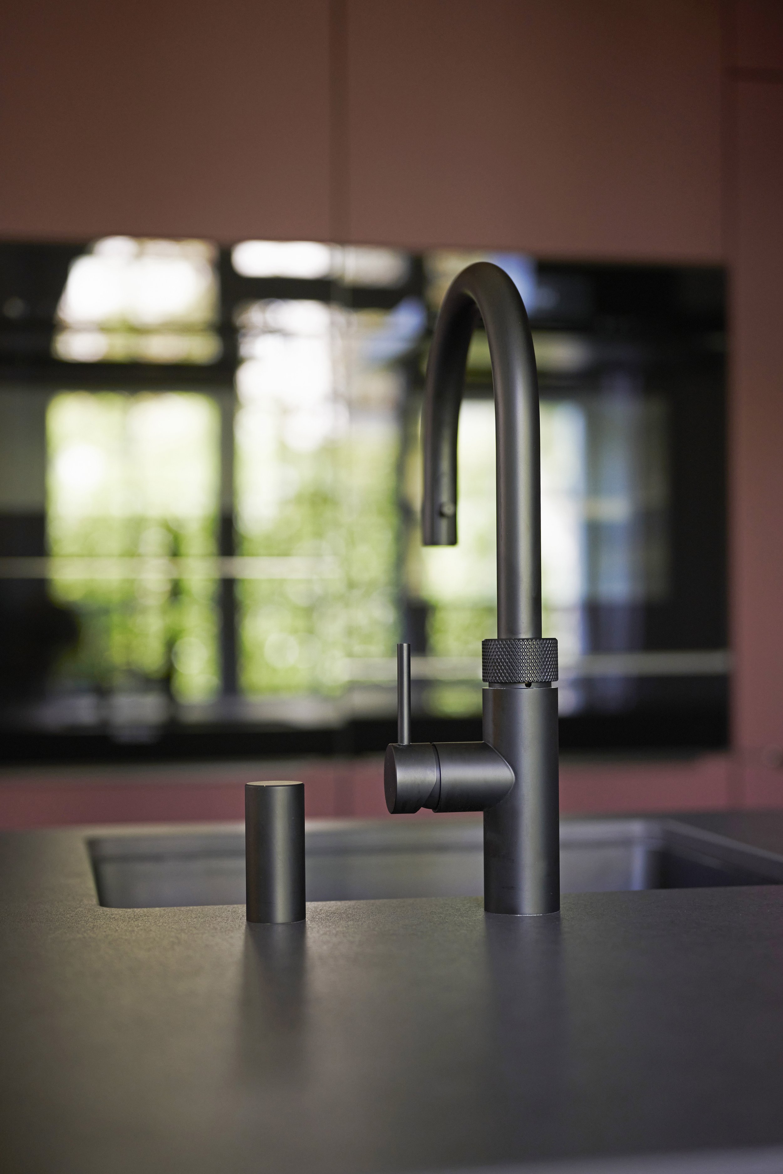 Moderne schwarze Küchenarmatur an einem schwarzen Küchenarbeitsplatten mit abgetrenntem Wasserhahn, im Hintergrund eine Fenster mit sichtbaren Bäumen und Gebäuden draußen.