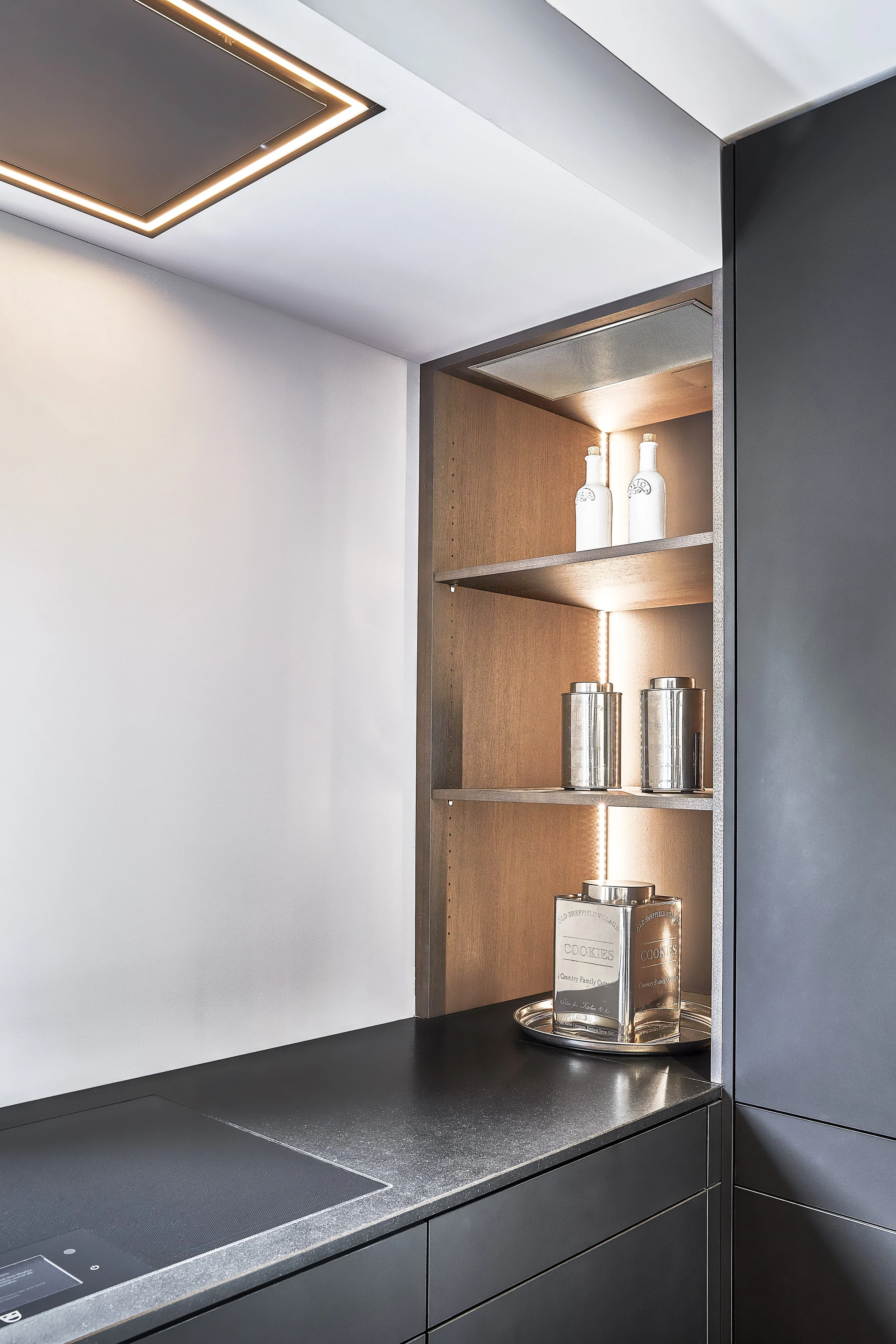 Moderne Küchenschrank mit Licht, offene Holzregale mit weißen Flaschen, metallene Behälter und eine Glasschüssel auf einem schwarzen Arbeitsplatten.