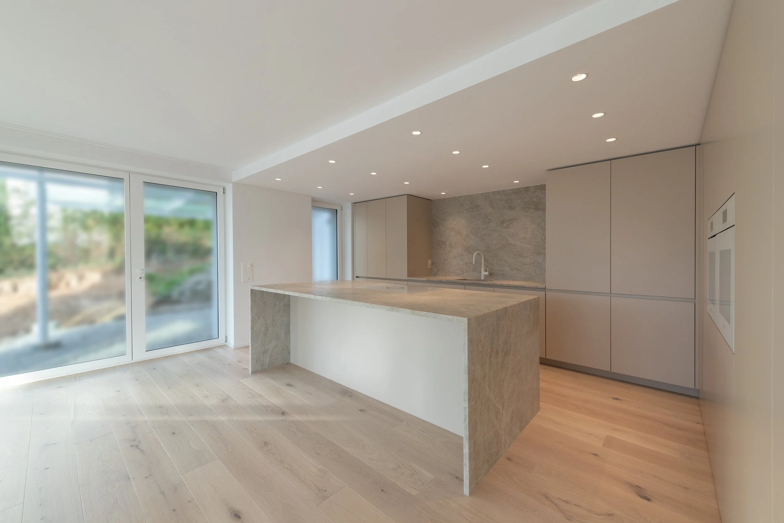 Moderne, minimalistische offene Küche mit einer Kücheninsel aus hellem Stein, grauen Einbaumöbeln und einem grauen Mauerakzent im Hintergrund, große Glasschiebetüren nach draußen, helles Holzparkett, dezente Hellraumleuchten an der Decke.