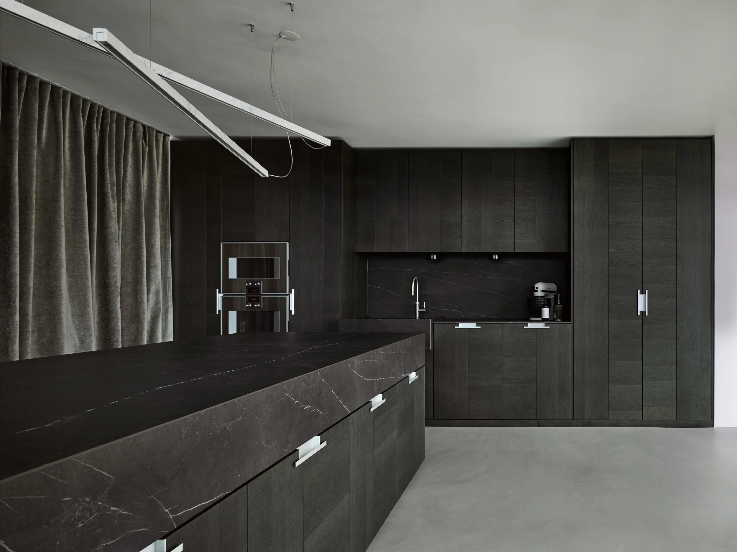 Moderne schwarze Küche mit schwarzen Schränken, schwarzer Arbeitsplatte, eingebautem Ofen, Dunstabzugshaube, Wasserhahn und Kaffeemaschine, beige Bodenfliesen und grauem Balkonvorhang.