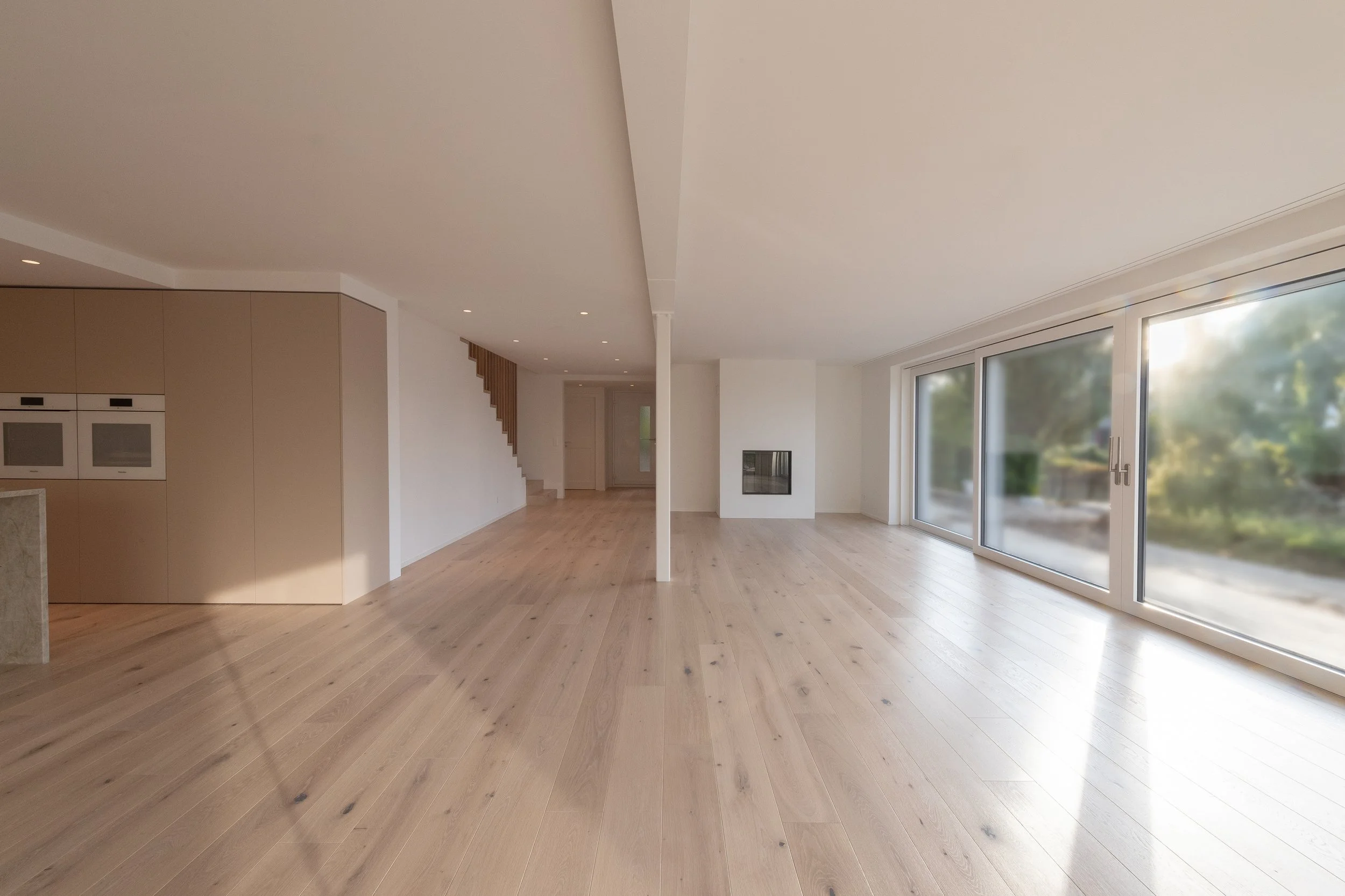 Ein großes, helles und minimalistisches Wohnzimmer mit Holzboden, großen Fenstern und einem modernen Kamin. Das Zimmer ist unmöbliert und bietet eine offene Raumgestaltung.