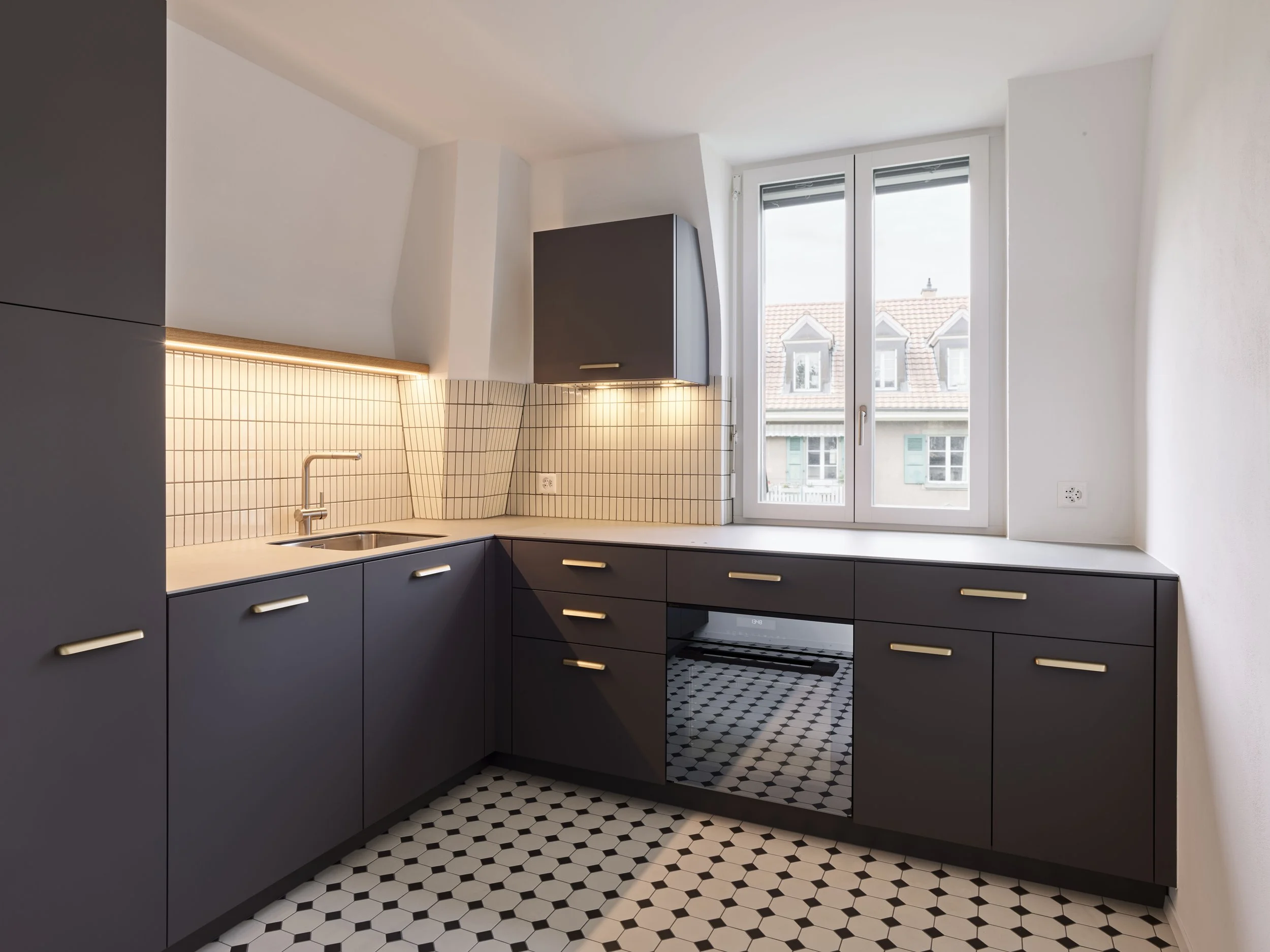 Moderne Küche mit dunklen Schränken, einer weißen Arbeitsfläche, einer Spüle, einem Herd und einem großen Fenster, das Blick auf Nachbarhäuser bietet.