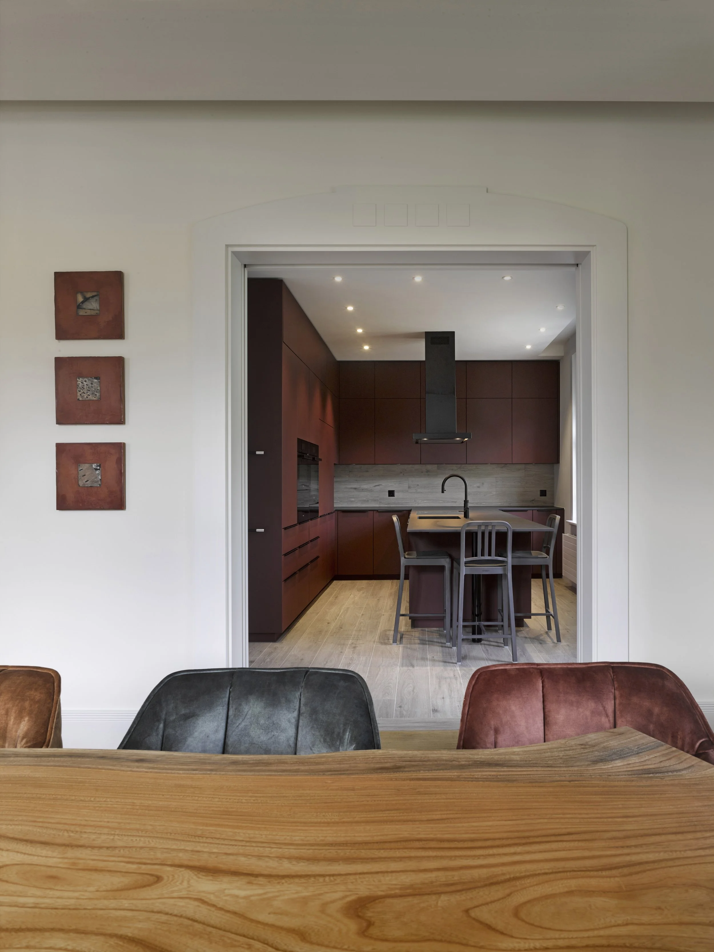 Blick durch einen Esszimmer in eine moderne Küche mit dunklen Schränken und einer Kücheninsel, von einem Holztisch aus fotografiert, mit Wanddekoration auf der linken Seite.